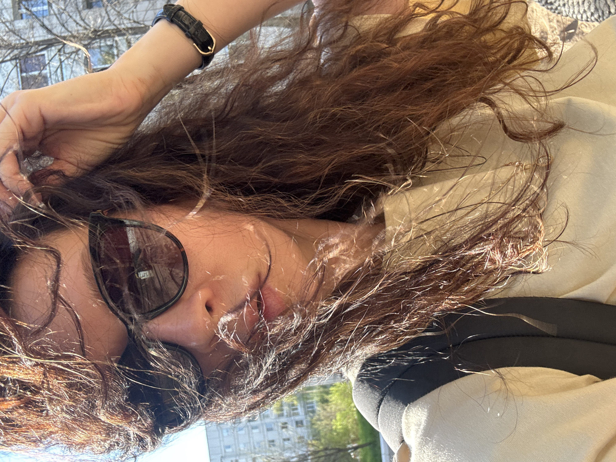 A woman is pictured outdoors in daylight. She has long, wavy brown hair, is wearing sunglasses, a light-colored jacket and a black wristwatch. In the background, a multi-story building and trees are visible, indicating an urban setting. Based on visual cues, it is not possible to confirm the specific city location as Chaikovsky, Russia without additional contextual information.