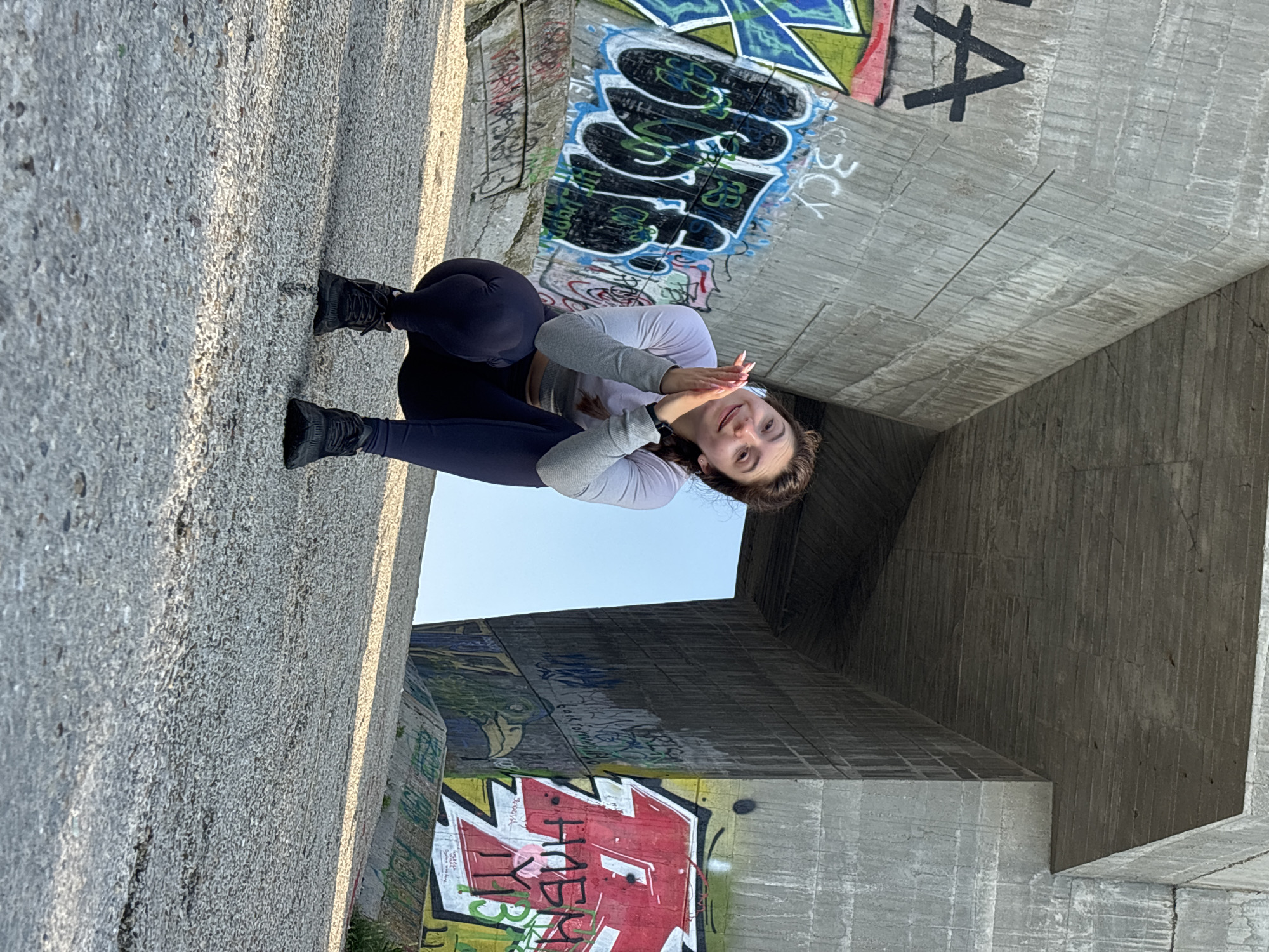 A young woman is captured in a full-body shot, crouching or sitting with her knees bent on a concrete surface. She is wearing a light-colored long-sleeved top, dark leggings, and dark athletic shoes. Her hands are clasped together near her chin, and she is looking directly at the camera with a gentle smile.
The setting appears to be beneath a large concrete overpass or bridge, characterized by massive, textured concrete structures. The ground and surrounding walls are made of this rough concrete. Many of the concrete surfaces, especially the walls and ceiling sections, are covered in colorful graffiti. Visible graffiti includes stylized letters and shapes in red, black, blue, green, and white. Specific legible elements include a large black "A", "BCY", and other complex letter designs.
Bright sunlight illuminates the scene, casting shadows on the ground, suggesting it is daytime, possibly late afternoon or early morning. A clear blue sky is visible through the openings between the concrete structures. The overall impression is urban and slightly industrial, with the vibrant graffiti contrasting with the monochromatic concrete. The location context is Varna, Bulgaria.