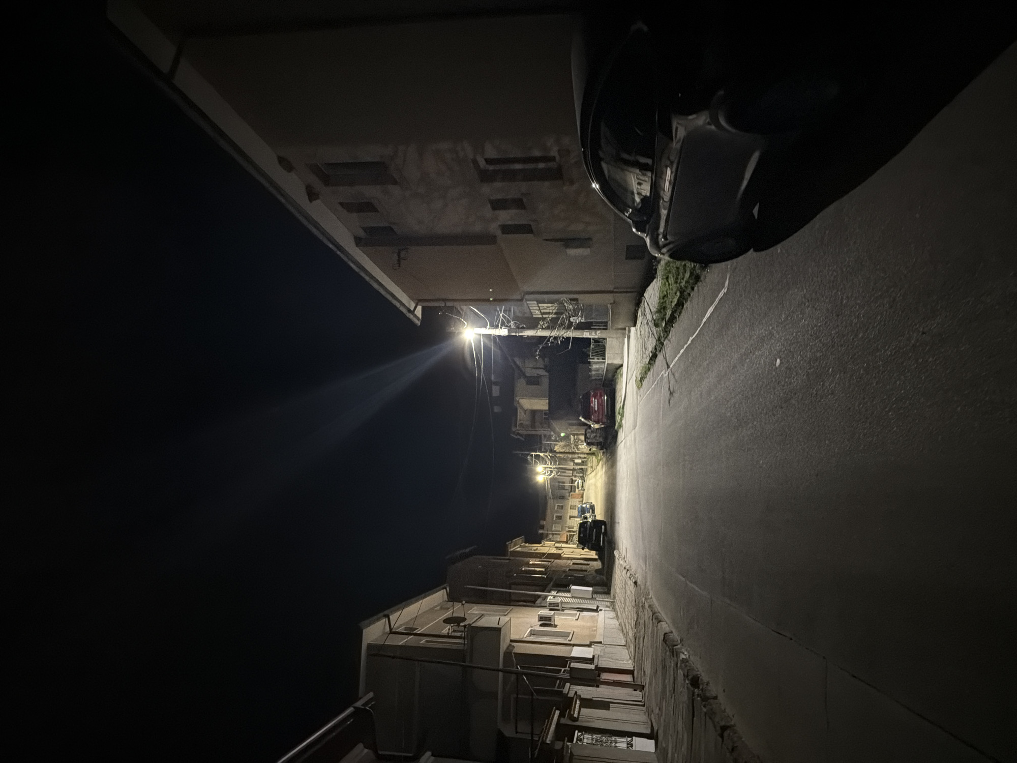 The image shows a narrow street at night in Panagyurishte, Bulgaria. The street is illuminated by streetlights, casting long shadows. Buildings line either side of the street, some with visible windows and balconies. A dark-colored car is partially visible on the upper right side of the frame. The sky is dark, suggesting a late hour. The overall scene is quiet and deserted.