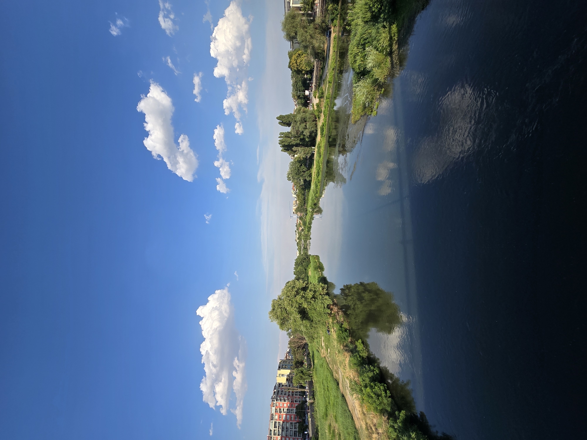 This media file is a vertical, landscape photograph capturing a wide, dark body of water reflecting a bright blue sky dotted with fluffy white clouds. The water is on the right side of the frame, while the sky occupies the left.
Along the left bank of the water, a lush green grassy embankment is visible, leading up to a line of trees and some buildings. The buildings are a mix of modern and older styles, with some appearing to be apartments or residential complexes, featuring red and yellow facades. Further back, more trees and some structures suggest a town or city. The trees are verdant, indicating it is likely summer or late spring.
On the opposite bank, closer to the center of the frame, more greenery and trees are present, with a pathway running alongside the water. A few small figures, possibly people, can be seen along this path, though they are not a prominent subject. The overall scene suggests a natural or semi-urban waterfront setting on a clear, sunny day, likely in the late morning or early afternoon given the bright light and clear sky. The reflection of the sky and clouds on the water is a notable detail, creating a symmetrical effect. There is no visible text in the image.