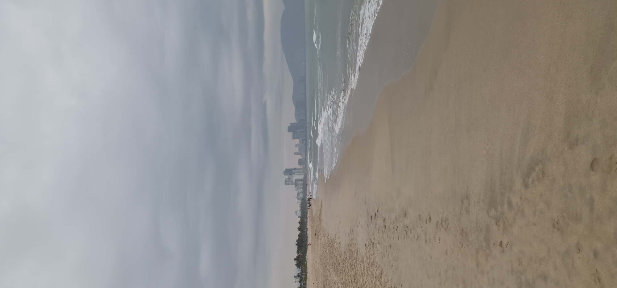 This wide-angle photograph captures a serene yet urban coastal scene in Đà Nẵng, Vietnam, under an overcast sky. The image is oriented horizontally, showcasing a long expanse of light brown sand in the foreground, marked with subtle ripples from receding waves and scattered footprints.
To the left, the gentle waves of a light green-grey ocean break onto the shore, leaving foamy white crests. The horizon features a continuous line of modern high-rise buildings, forming a city skyline that stretches along the coastline. These structures appear somewhat hazy, blending into the muted tones of the sky. Behind the city, rolling mountains rise, their silhouettes partially obscured by the heavy, uniform grey cloud cover that dominates the upper portion of the frame. A thin strip of palm trees or similar coastal vegetation runs along the edge of the beach, separating the sand from the urban development.
Several small figures of people are visible on the beach, mostly near the water's edge, engaged in casual strolls. The weather appears cloudy, providing diffused, soft lighting across the entire scene without harsh shadows. The overall mood is calm and somewhat subdued, reflecting a non-sunny day at a busy coastal resort city. There is no visible text in the image.