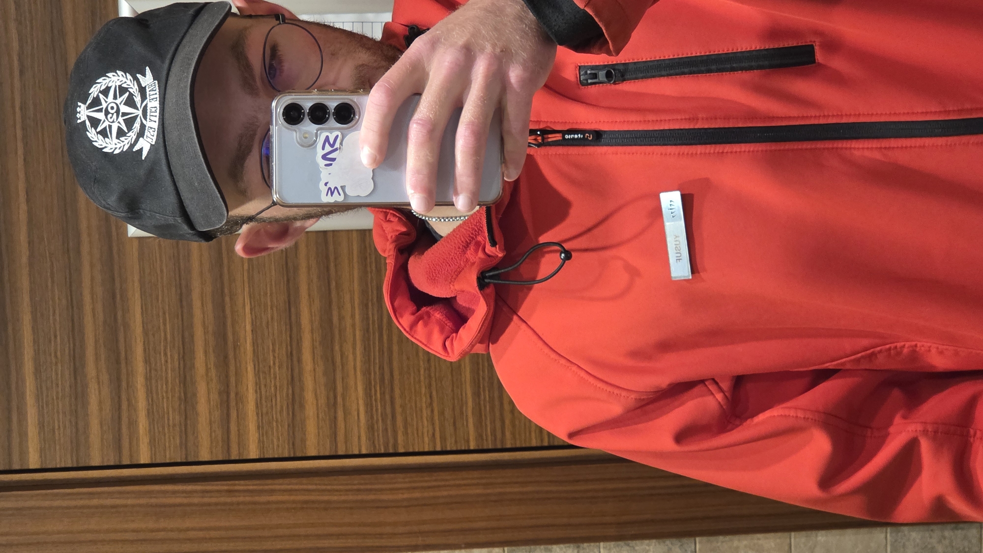 A man is taking a selfie in front of a wooden wall. He is wearing a black baseball cap with a compass logo, glasses, and a red jacket. He is holding a smartphone in his left hand. The phone has a clear case with stickers. The jacket has black zippers and a reflective tag. The location is Unknown, Unknown.
