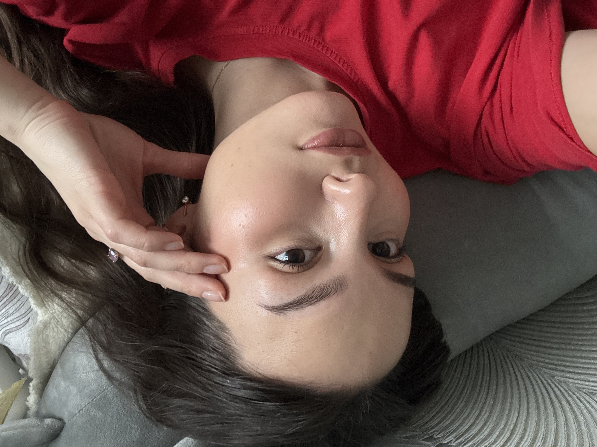 A young woman with dark hair and fair skin is lying down on a gray patterned surface, possibly a pillow or blanket. She is wearing a red t-shirt. Her head is tilted, and she is looking directly at the camera. Her right hand is near her face. She appears to be indoors. There is no evidence in the image to suggest this image was taken in the city of Chaikovsky, Russia.