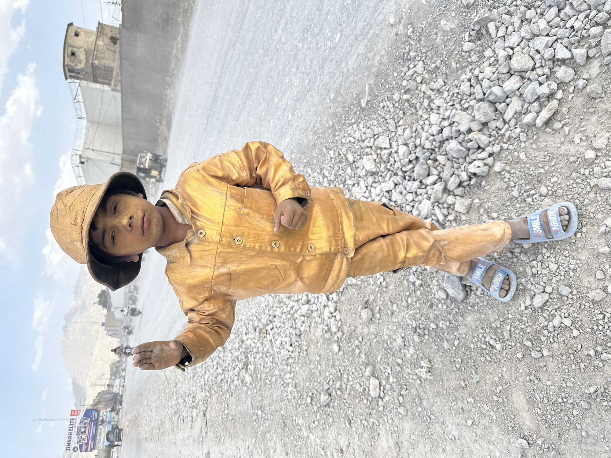 A young boy is lying on a gravel road, oriented towards the viewer. He is wearing a tan-colored hat, a button-down shirt, pants, and blue sandals. His face and clothes are covered in dust. The background consists of a city street scene with vehicles and buildings. In the distance, there is a mountain range. The location appears to be Quetta City Tehsil, Pakistan.