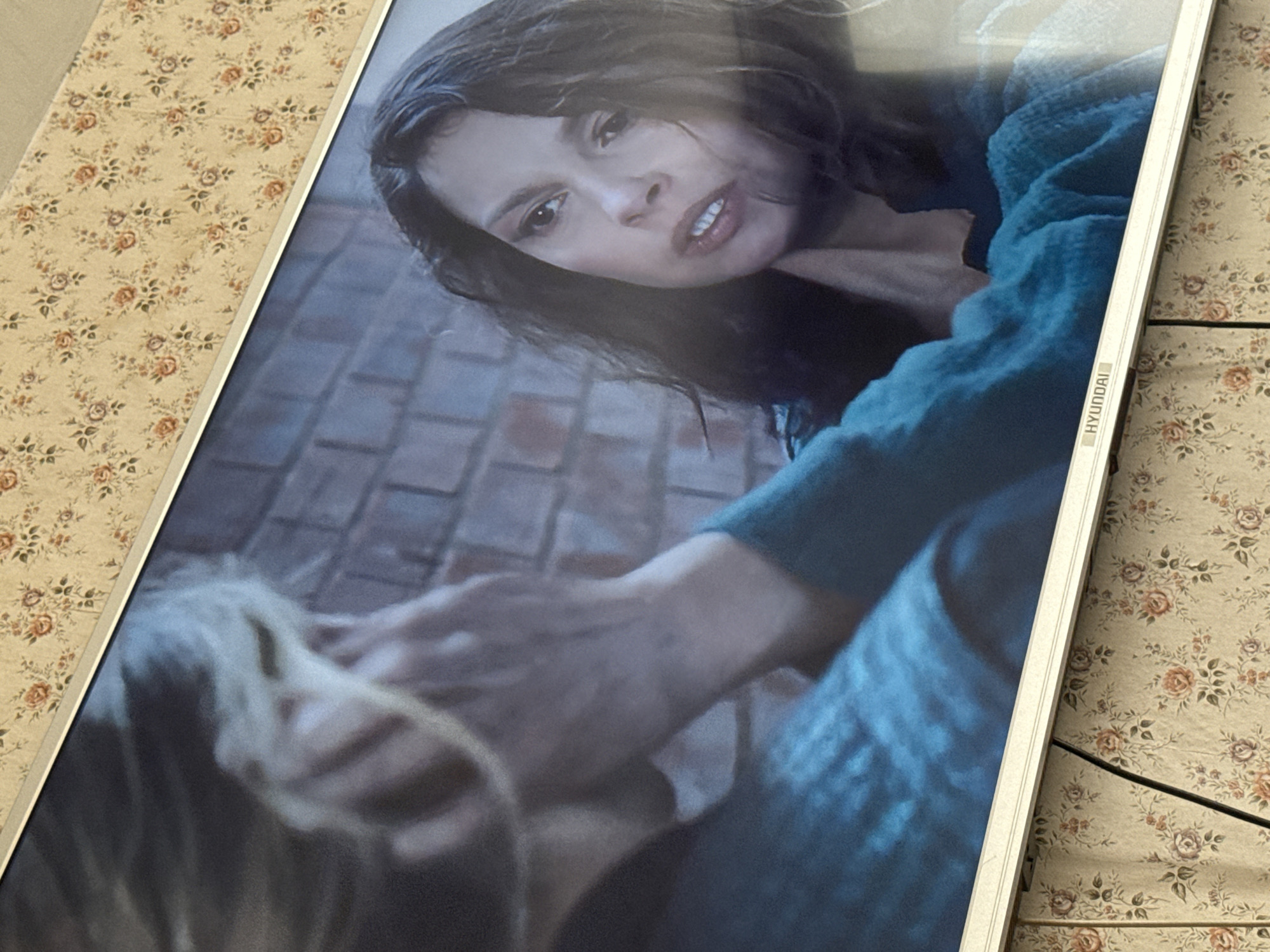 The image shows a close-up shot of a Hyundai television screen displaying a scene from a film or television show. The scene depicts a woman with dark hair, wearing a blue garment, looking down intensely at another person with lighter hair, who appears to be lying on the ground. The background suggests a brick or stone surface. The television is placed on a surface covered in a floral-patterned fabric. There is no information to suggest that this image was taken in the city of Mytishchi, Russia.