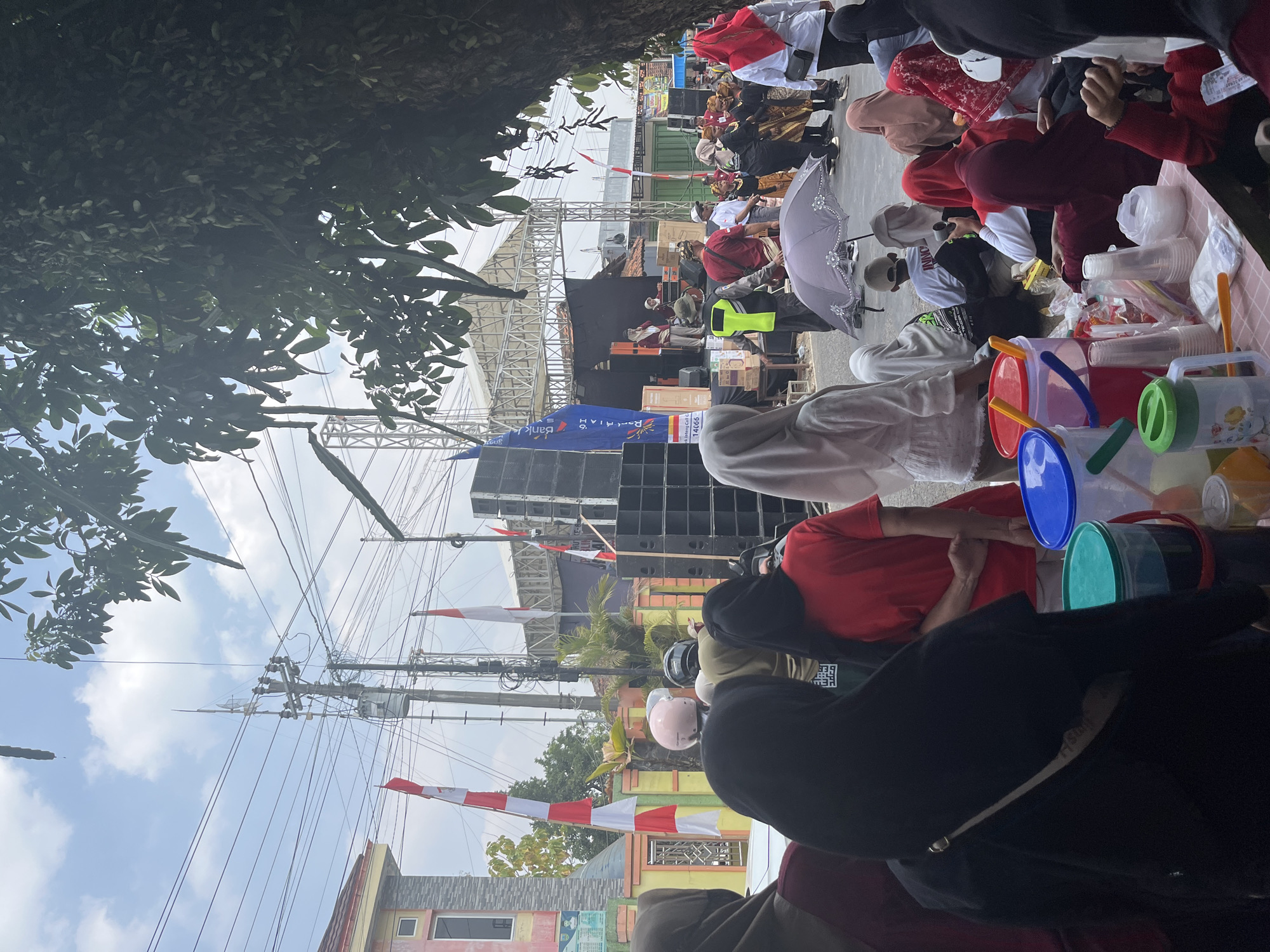 The image depicts an outdoor event in Glagah Kulon, Indonesia, with a crowd gathered around a stage. A large sound system is set up to the right of the stage, and there are flags and banners visible. Several people are standing and sitting, some wearing traditional clothing. The weather appears to be overcast. On the left side of the image, there is a building with a red roof. The foreground shows a table laden with drinks and food items. People are engaged in various activities, including observing the stage, conversing, and preparing food. The overall scene suggests a community gathering or celebration.