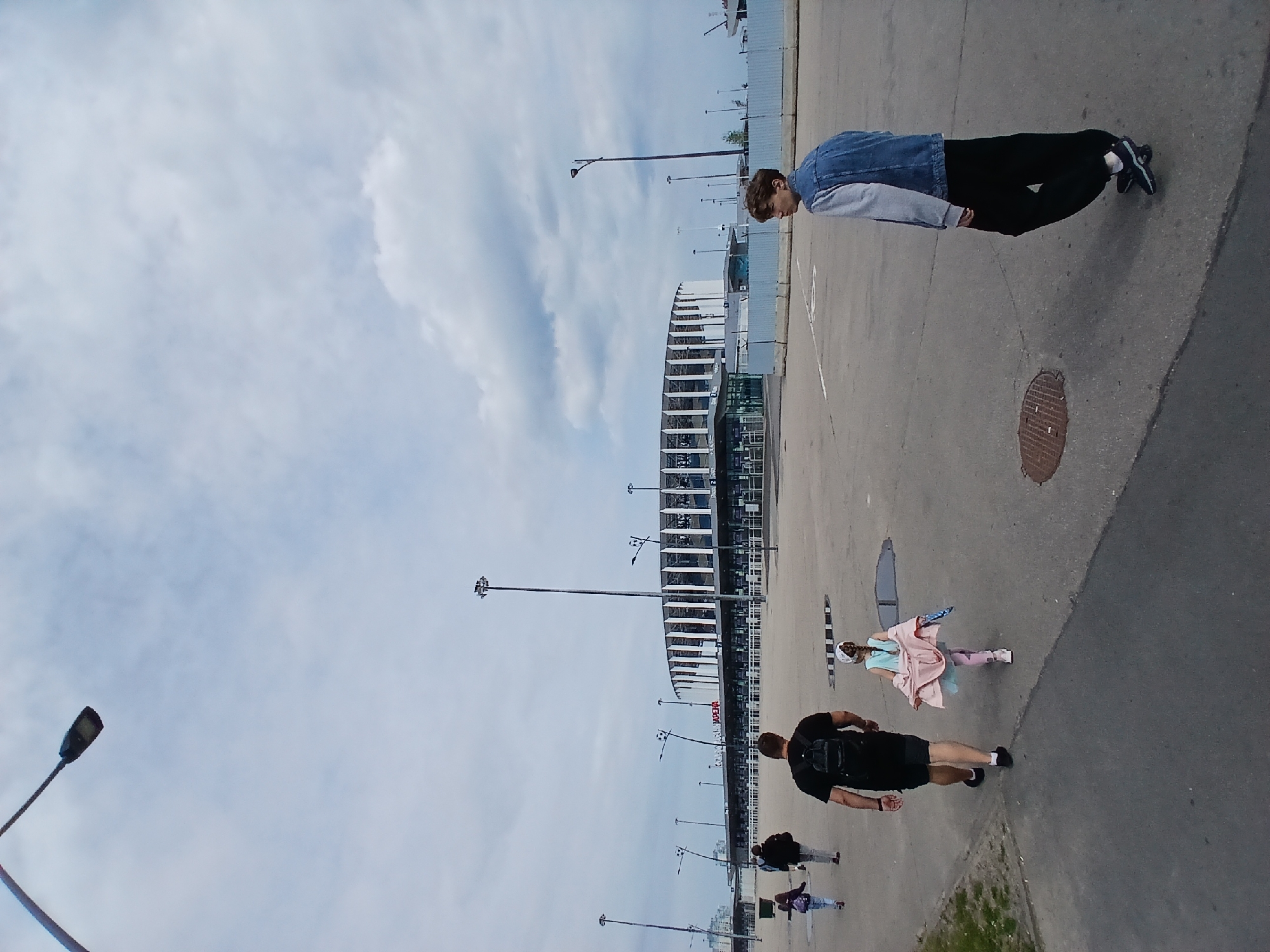 A group of people are walking on a paved area in front of a large, circular stadium with a white, ribbed exterior and a dark glass facade. The stadium has signs that appear to read "SOKOL ARENA". In the foreground, a man wearing a black t-shirt and shorts with a backpack walks away from the camera. To his right, a young girl in a pink dress and leggings walks in the same direction. Further to the right and closer to the camera, a teenage boy wearing a denim jacket over a grey hoodie and black pants walks away from the camera, looking over his right shoulder. In the background, a few other individuals are also walking away from the camera, towards the stadium. The sky is overcast with some clouds. The scene appears to be in an open area, possibly a plaza or walkway leading to the stadium. The location is identified as Nizhny Novgorod, Russia.