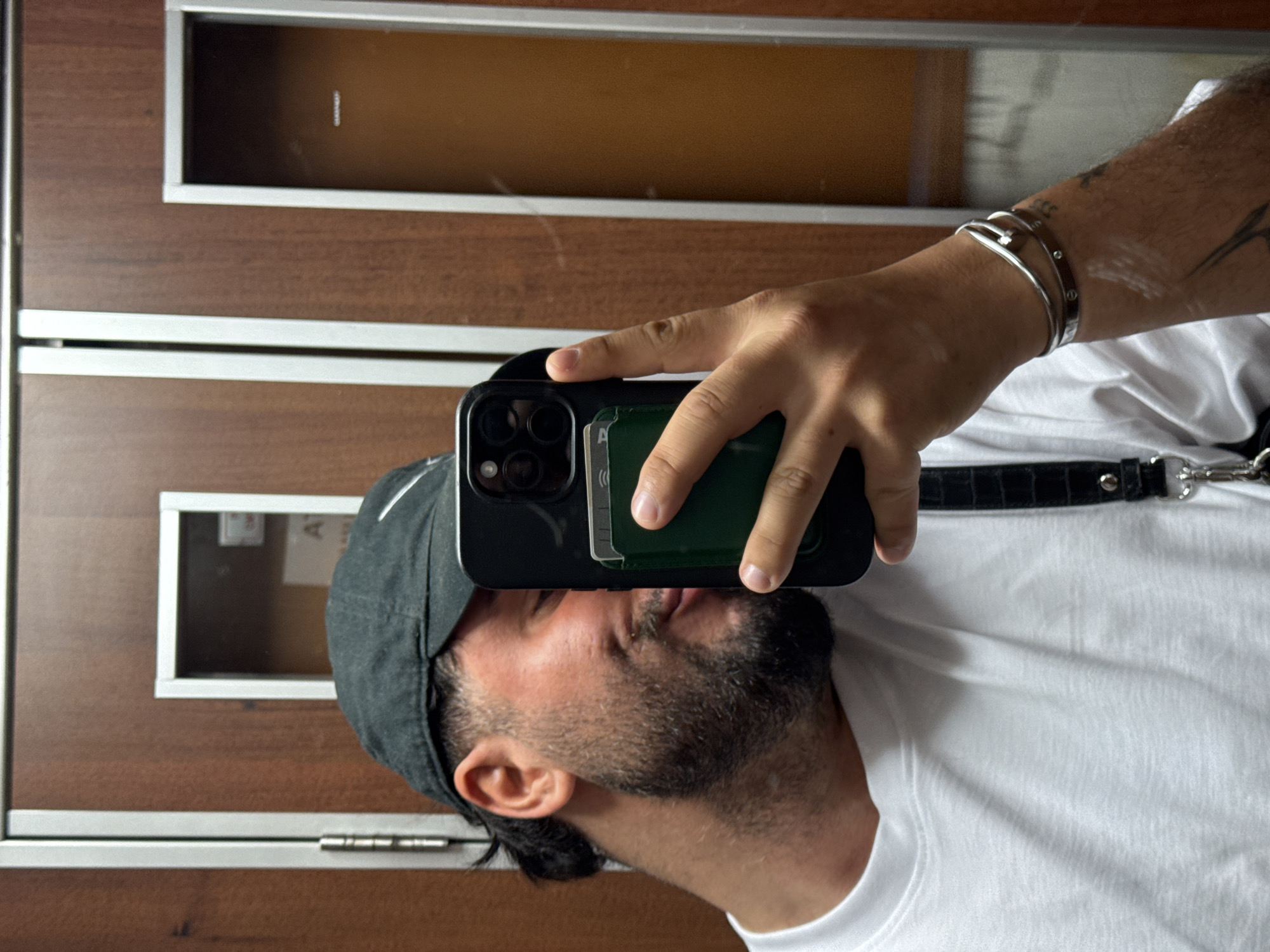 A man is taking a selfie in front of a brown door in Milan, Italy. The door has rectangular window panes and silver framing. The man is wearing a dark cap and a white t-shirt. He has a beard and is holding a black smartphone with a green wallet attached to the back. He's wearing silver bracelets on his left wrist. Part of a black strap is visible over his shoulder.