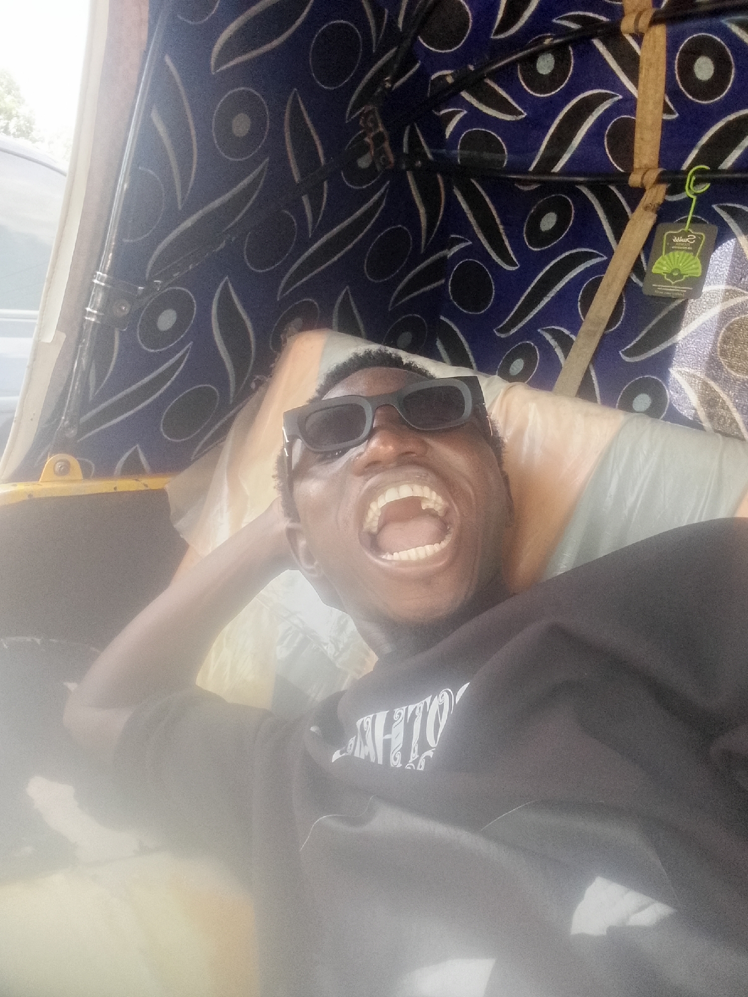 A man with dark skin and short, dark hair, wearing black sunglasses and a dark t-shirt with a white design, is lying down inside what appears to be an auto rickshaw or similar vehicle. The vehicle's interior has a patterned blue and white roof. The man has his head resting on a light-colored pillow or cushion and his mouth is wide open in a laugh or shout. The scene appears to be Bukuru, Nigeria.
