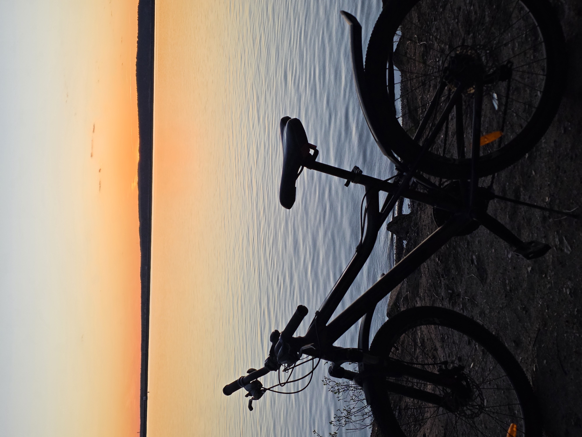 A bicycle, likely a mountain bike given the tire size and suspension visible, is positioned along the embankment of a body of water, presumed to be the Kama River based on the Chaikovsky, Russia location. The bike is heavily silhouetted, making details difficult to discern, but the general outline of the frame, wheels, seat, and handlebars are visible. The background suggests a sunset or sunrise over the water. The ground is a rough, earthy surface. No people are visible. The scene evokes a sense of quiet solitude at dawn or dusk.
