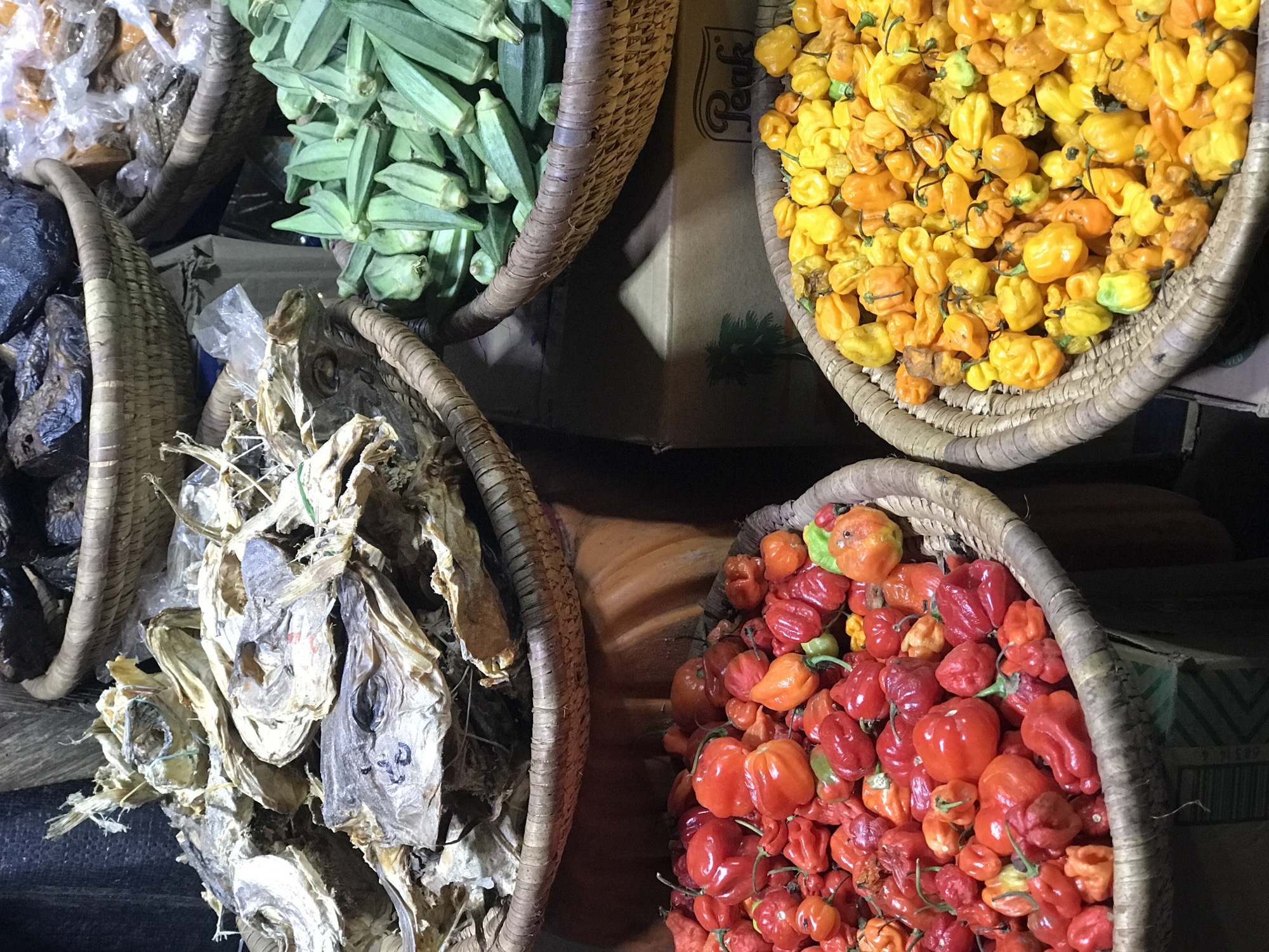 The image captures a vibrant market scene in Abuja, Nigeria. Four woven baskets are prominently displayed, each filled with different food items. The top left basket contains dried goods of an unidentified type. The top center basket is filled with green okra pods. The top right basket contains a pile of yellow and orange scotch bonnet peppers. The bottom right basket is filled with red scotch bonnet peppers. The bottom left basket contains dried fish, including whole dried fish heads. The baskets are sitting on or next to a cardboard box with an image of a palm tree and the word "Peak" on it. The lighting in the scene is somewhat dim. The scene depicts a common market offering of food staples found in Nigeria.