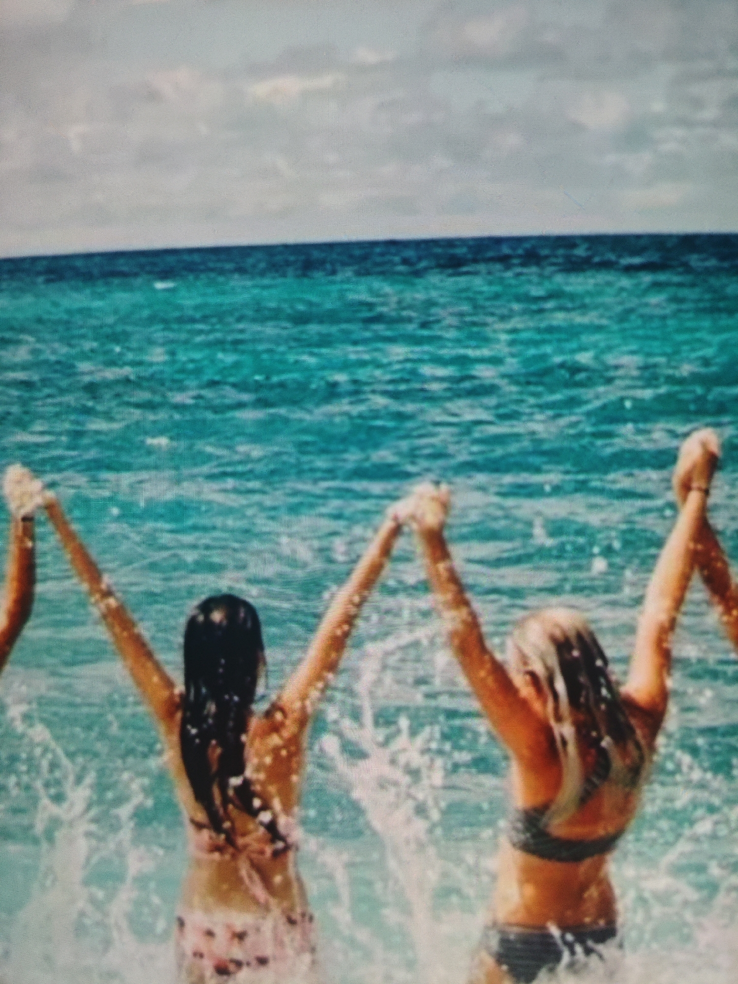 The image shows a group of women from behind, standing in the ocean and raising their arms. The water is a vibrant turquoise color, and the sky is overcast with some clouds. The women are wearing swimsuits and appear to be having fun as they splash in the water. One woman has long dark hair, while another has blonde hair. The scene is likely a beach or coastal area. The location is not specified as being in Krasnaya Polyana, Russia.