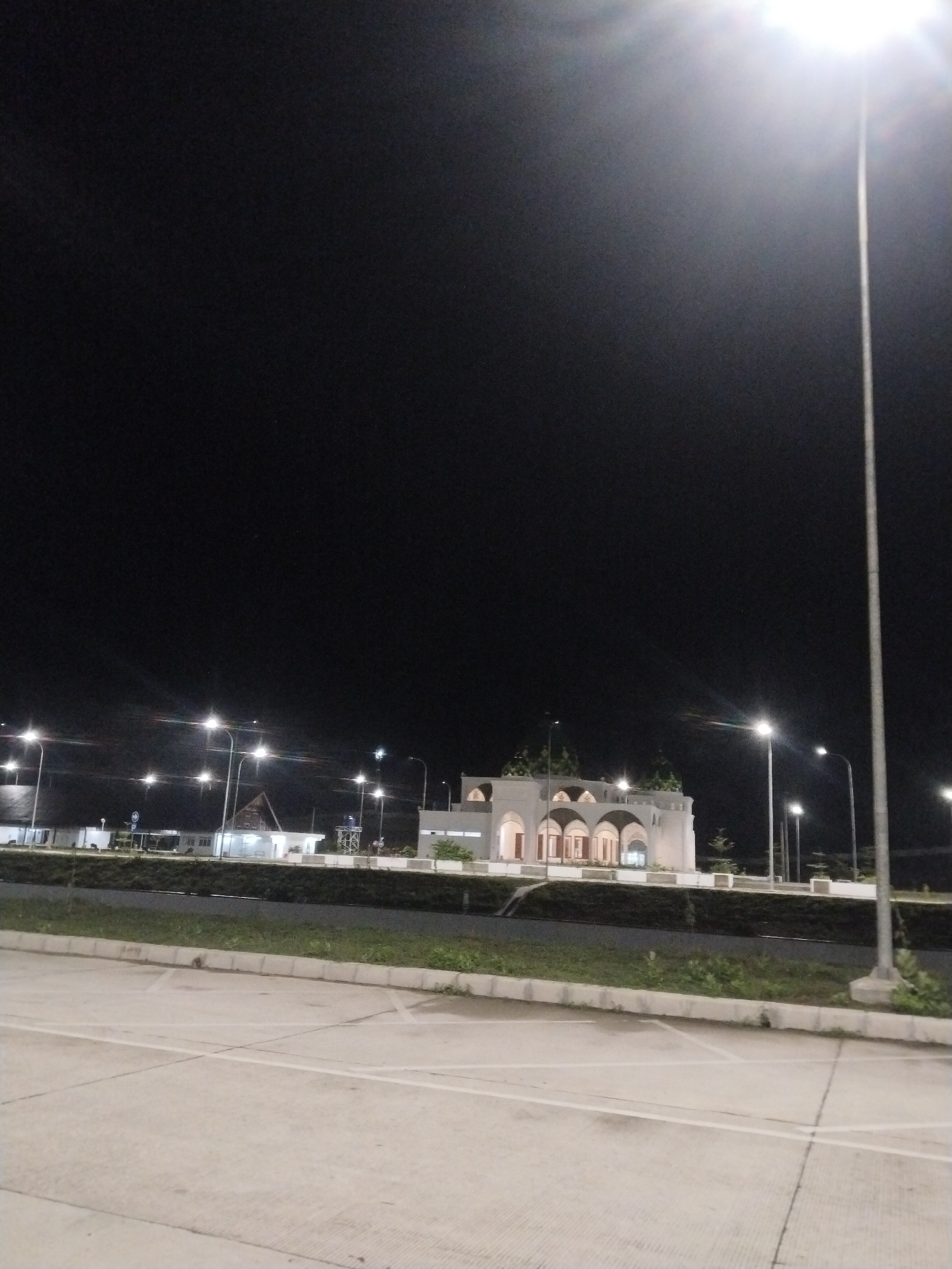 The image depicts an outdoor scene at night, likely in Keureuweungkrueng, Indonesia, given the architectural style of the building. A brightly lit parking lot occupies the foreground, with white parking lines painted on the surface. Beyond the parking lot is a strip of greenery with low plants. Further back is a row of buildings illuminated by streetlights. The buildings appear to be a mix of governmental or public structures, including a distinctive white building with arches that may be a mosque or community center. Tall streetlights are visible throughout the scene, casting a bright glow. The sky is dark and lacks detail. There are no people visible in the image.