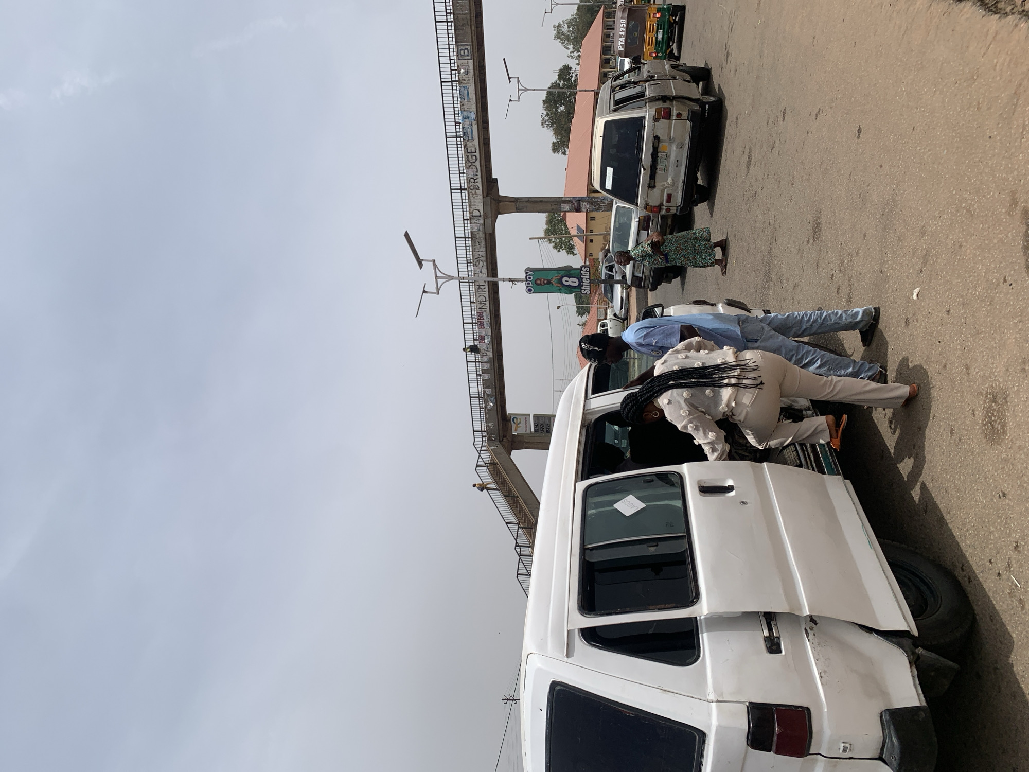 The image captures a vibrant street scene, likely in an urban or semi-urban area, under a bright, somewhat hazy daytime sky. The primary focus is on a white passenger van where several individuals are engaged in activity. A woman, dressed in a white patterned top and light-colored trousers, is seen stepping into the side door of the van, while a man in a blue shirt and jeans stands beside her, his attention directed towards the van's interior.
In the immediate foreground, the rear of the white van shows some signs of use. To the left, further down the road, another white vehicle is parked, and a woman in a green patterned dress stands on the roadside. In the background, part of a yellowish-green tricycle, commonly known as a Keke NAPEP in Nigeria, is visible behind another parked car.
A prominent feature in the mid-ground is a concrete pedestrian bridge with dark metal railings spanning across the road. Text inscribed on the bridge structure reads "ROAD BRIDGE" and "ANDRI." Beneath the bridge, an advertising board displays "OPay" and "8 Shields," accompanied by an image of a person. Various buildings with red roofs can be seen lining the street in the background. The road surface is asphalt, with scattered debris. On a rear side window of the foreground van, a sticker clearly displays the word "LAGOS." The license plate of a white vehicle in the background reads "PTA 375B."