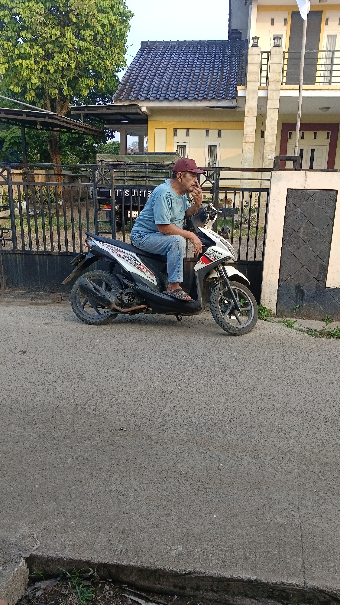 In Parung, Indonesia, an elderly man wearing a light blue t-shirt, blue jeans, and a maroon baseball cap is sitting on a white and black Honda Beat scooter. He is facing to the left, with his left hand near his face, appearing to be touching his chin or mouth. The scooter is parked on an asphalt road, with a black metal fence behind it. Beyond the fence, a multi-story building with yellow and white walls and a dark grey tiled roof is visible. A large tree with green leaves stands to the left of the building. A truck with "MITSUBISHI" written on its side is partially visible behind the fence. The overall scene depicts a quiet street in a residential area.
