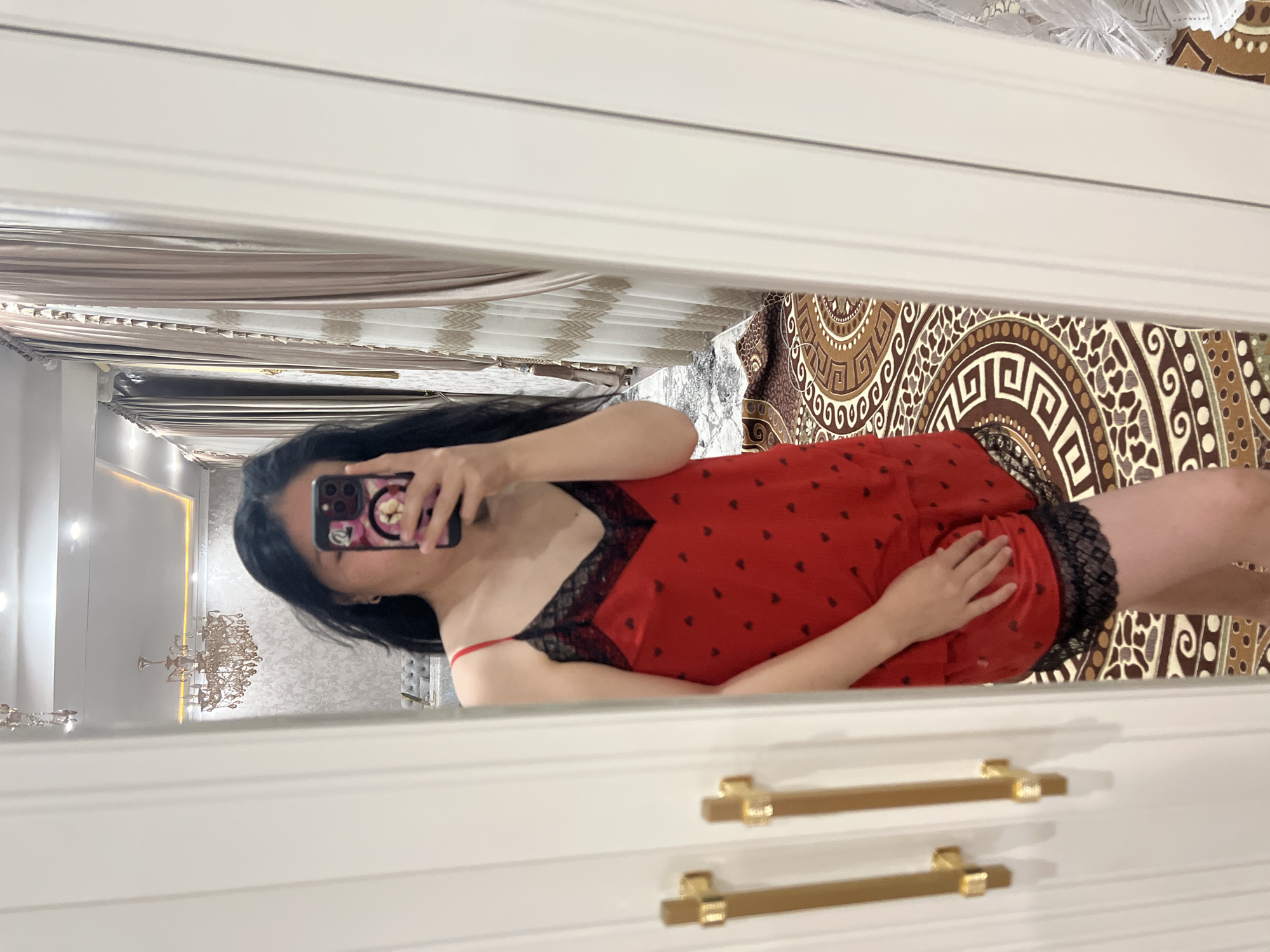 A young woman with long, dark hair takes a mirror selfie in a room. She is wearing a red slip with a black lace trim and a pattern of small black hearts. The slip has thin spaghetti straps. Her right hand is resting on her hip, and she is holding a smartphone in her left hand, which is partially obscuring her face. The mirror reflects a chandelier hanging from the ceiling, ornate curtains, and a patterned rug on the floor. The woman is standing in front of a white wardrobe with gold handles. The room appears to be a bedroom or dressing area, with a luxurious and possibly opulent decor. There is no indication that this image was taken in Kara-Dıykan, Kyrgyzstan.