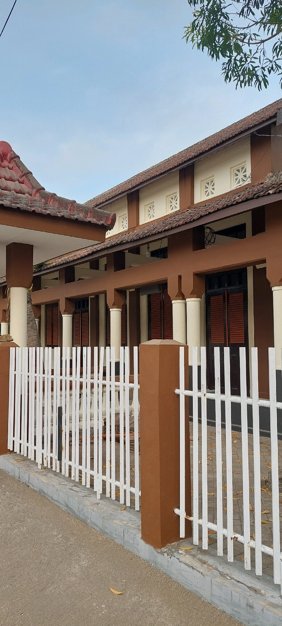The image shows the exterior of a one-story building with a brown facade and a tiled roof. The building has several large white columns supporting a covered walkway. There are windows with decorative patterned vents above the walkway. A white picket fence runs along the front of the property, and a concrete sidewalk is visible in the foreground. The sky is overcast. The image is taken from a ground-level perspective.