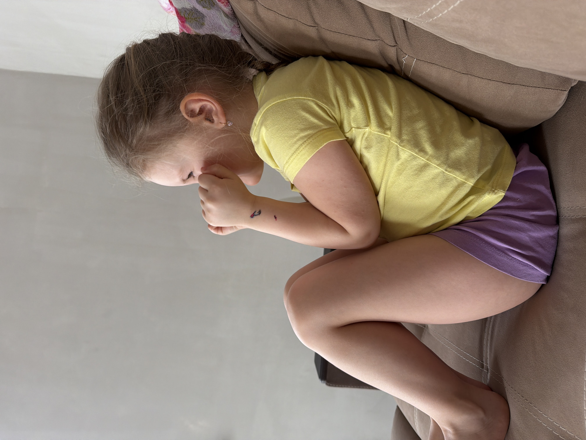 A young child is curled on a brown couch. The child wears a yellow short-sleeved shirt and purple shorts. The child's face is obscured as they appear to be holding their hand to their mouth. The child is indoors, with a gray wall visible. The location is inferred as Chaikovsky, Russia, based on external information. Details suggest a domestic setting.