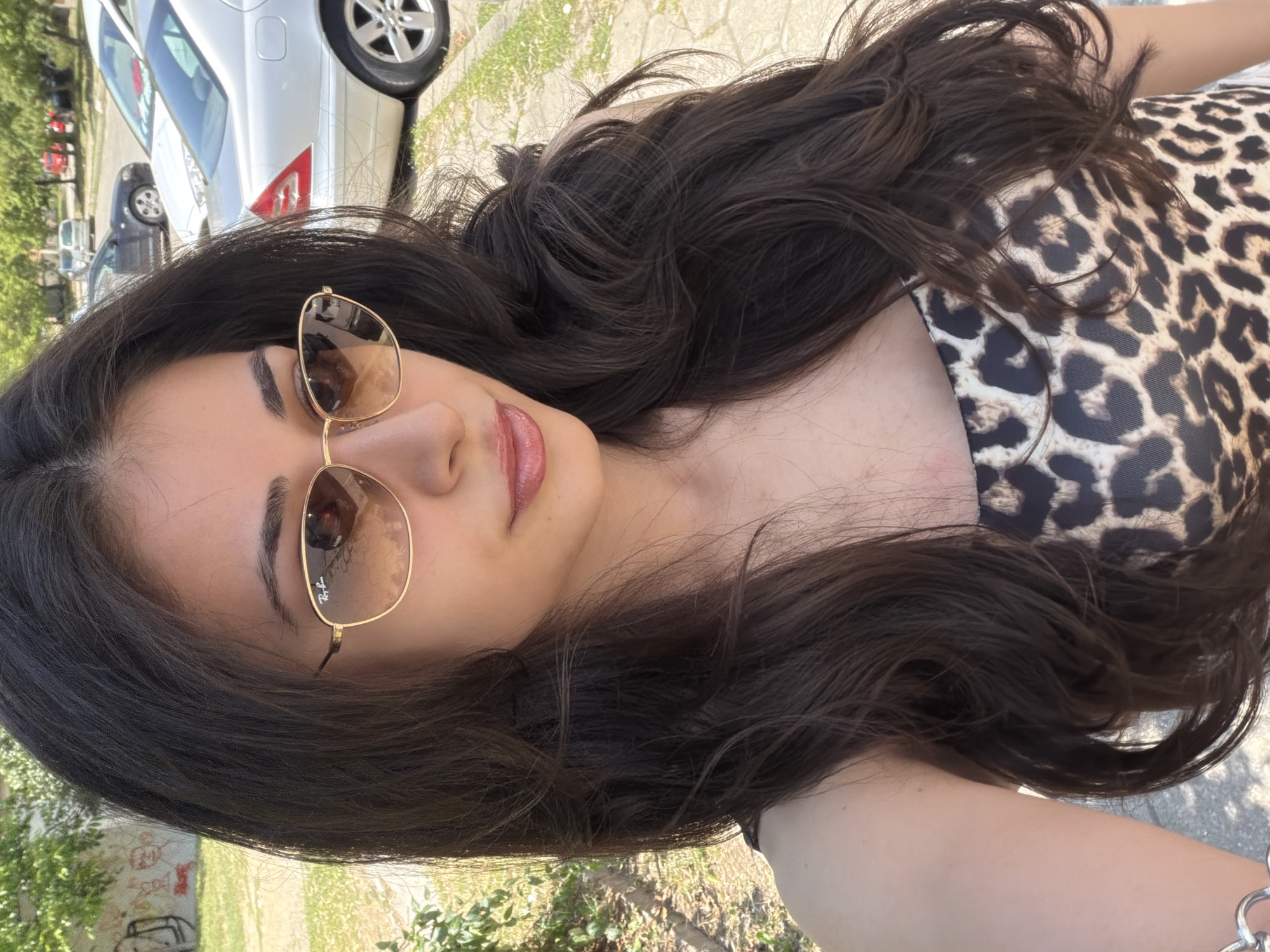 A medium shot depicts a woman with long, dark hair wearing brown-tinted Ray-Ban sunglasses and a leopard-print top. The background reveals a city scene with cars parked alongside a street. The location appears to be Blagoevgrad, Bulgaria, based on the architectural style and vegetation. The woman is the central subject, possibly taking a selfie.