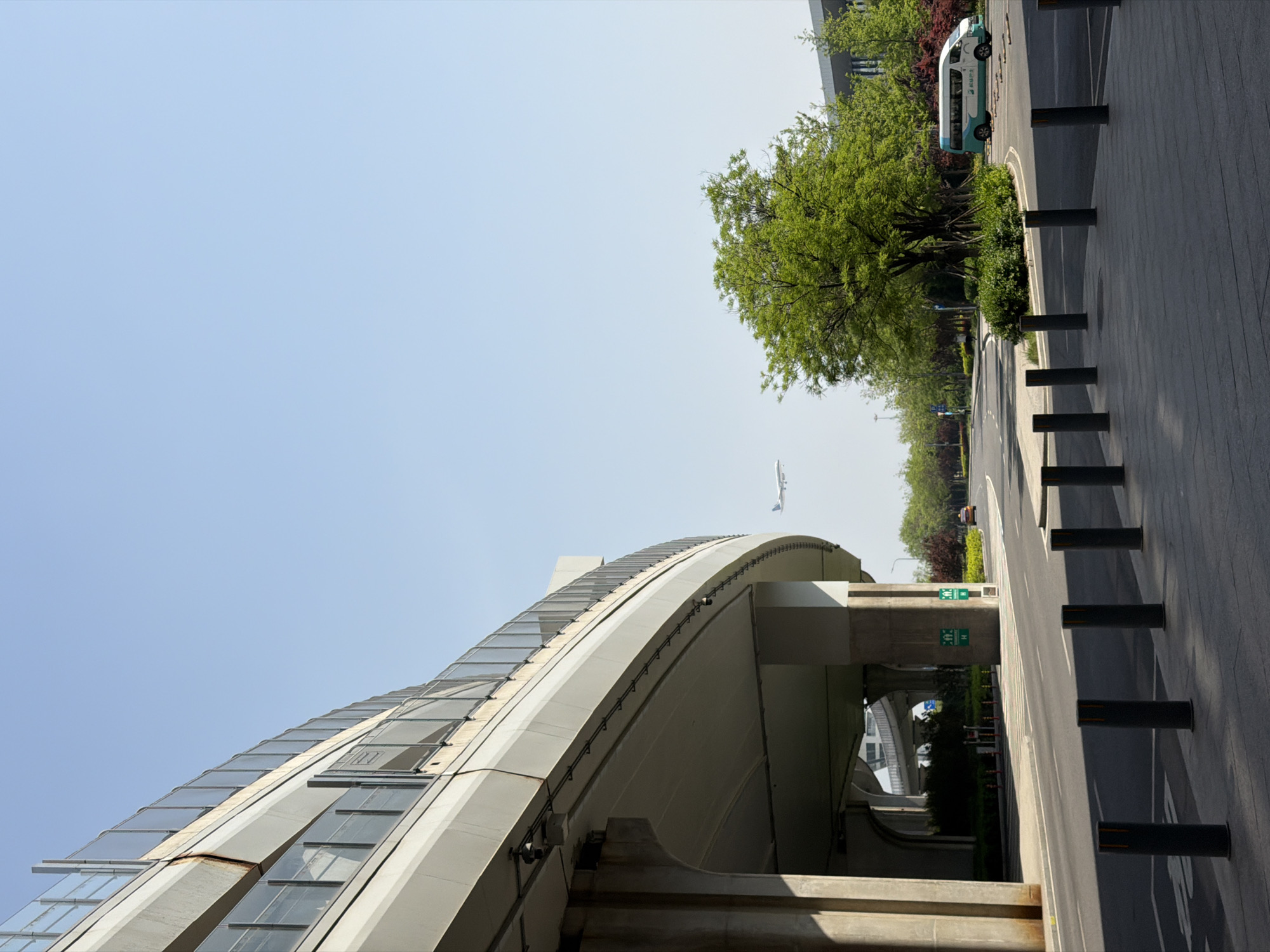 The image shows a street view in Langfang, China. A curved building with glass windows is on the left side of the image. Above the building in the sky, an airplane is flying. A road with black bollards lines the right side of the image. A line of trees with green leaves runs parallel to the road. Further down the road, a white van is visible. A pedestrian is standing near the trees. In the background, more buildings can be seen under an overpass.