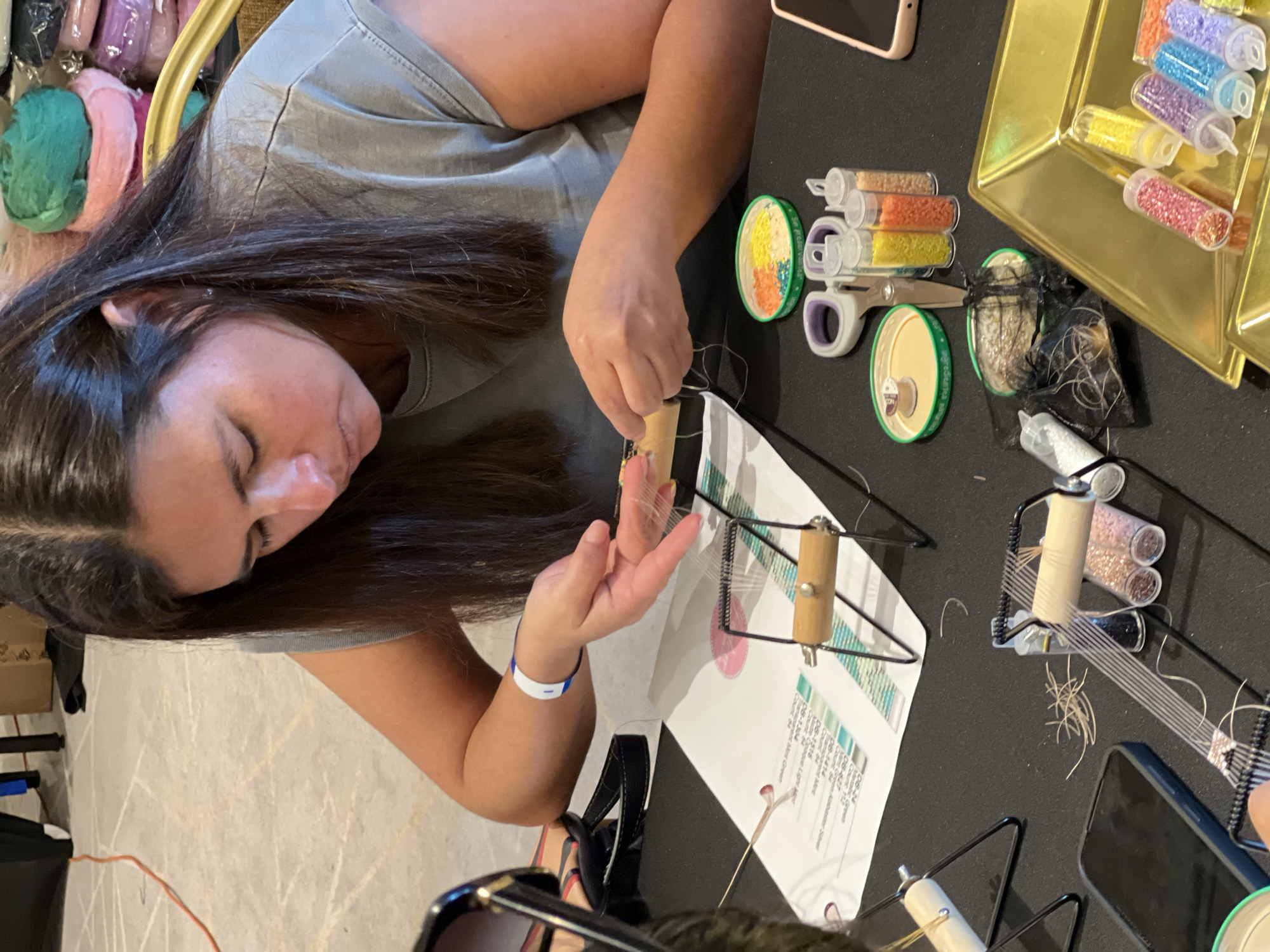 Arica, Chile: A medium shot, oriented vertically, shows a woman with long, dark hair working with a small weaving frame on a dark surface. The woman is wearing a gray t-shirt and has a white wristband on her left arm. She is focused on the weaving process, with her hands manipulating the thread. Around the weaving frame are various supplies, including tubes of colorful beads, spools of thread, and scissors. A golden tray holding more tubes of beads is visible in the upper right corner. A piece of paper with color swatches and text is also on the table. There are two mobile devices. The background is indistinct, but there are bags filled with colorful thread.