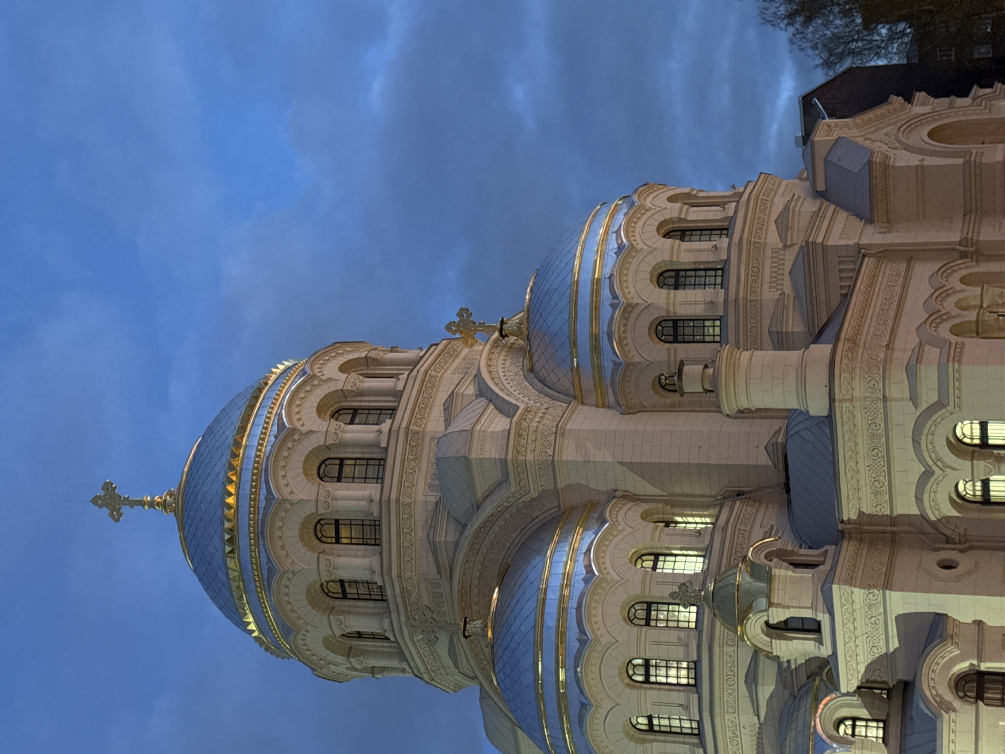 An illuminated Eastern Orthodox cathedral dominates the frame, captured from a low angle looking upwards, creating a majestic perspective. The building features multiple prominent blue domes, accented with gleaming gold bands and topped with golden Orthodox crosses. The largest dome, positioned centrally, is particularly striking with its scale and intricate detailing.
The cathedral's facade is constructed from a light-colored material, likely stone, and is richly decorated with elaborate architectural elements. These include arched windows with dark grilles, decorative columns, intricate carvings, and patterned reliefs. The windows emit a warm, yellow light, suggesting the interior is lit and contrasting with the cooler exterior illumination on the building. The intricate details of the arches and cornices are highlighted by this lighting.
The sky above is a deep, dark blue, indicating either dusk or nighttime, with soft, indistinct cloud formations. The artificial lighting on the cathedral casts soft shadows and emphasizes its ornate structure against the dim sky. In the upper right corner, parts of dark, bare tree branches and a darker, less distinct building are visible, hinting at the surrounding urban environment.
No people or explicit activities are visible, focusing solely on the architectural grandeur of the structure. The overall impression is one of solemn beauty and historical significance. The building is the Naval Cathedral of St. Nicholas in Kronstadt, Saint Petersburg, Russia. There is no visible text in the image.