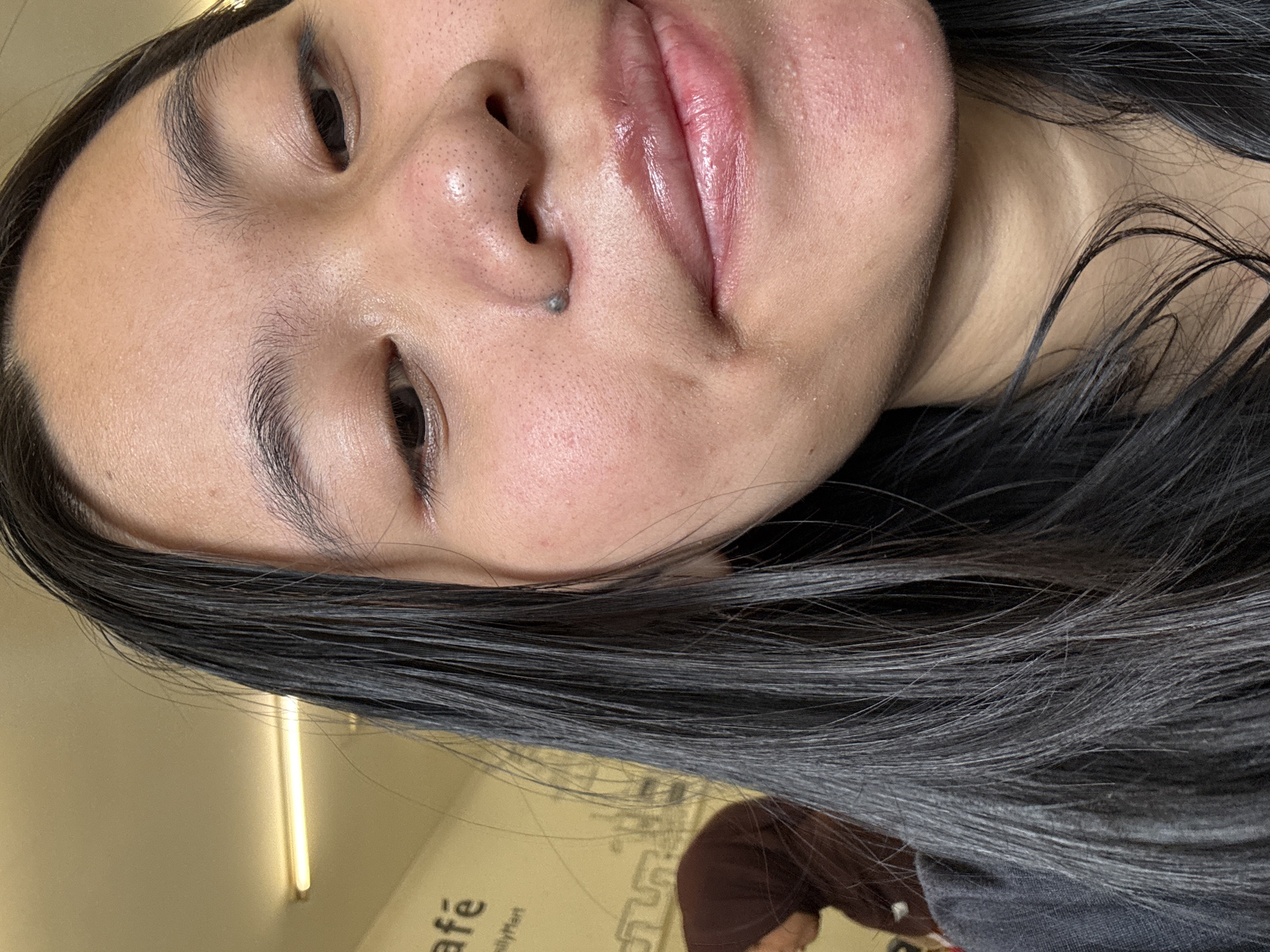 A close-up, selfie-style image features a young woman with dark hair in a dimly lit indoor environment. Her face, lit from above, is the primary focus, showing skin with visible pores and some redness. She has dark eyebrows and dark eyes, with her lips appearing glossy and slightly parted. A small, dark mole is visible on the bridge of her nose. She is wearing a dark shirt. In the background, a blurred figure wearing a brown hijab is visible, and text that appears to read "café" and "lyMart" is present on a wall. A fluorescent light fixture is also visible on the ceiling. The overall scene suggests a casual setting, possibly a cafe in East Jakarta, Indonesia, given the context.