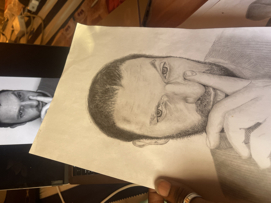 A dark-skinned hand, wearing a silver-toned ring on the fourth finger, holds a pencil drawing of a male portrait in the foreground. The drawing, rendered in graphite on light-toned paper, depicts a male subject with short, dark hair and a beard. The subject's right index finger is pressed against the right side of his nose, with his right thumb positioned beneath his chin. His gaze is directed forward.
In the upper-middle background, an open laptop screen displays a monochrome photograph of the identical male subject in the same pose, serving as the reference image for the drawing. The laptop's keyboard is partially visible beneath the screen. To the right of the laptop, blurry objects and cables, including a white electronic device, are visible on a dark surface, illuminated by warm ambient light.
There are no discernible visual elements within the image to specify a location in the city of Jos, Nigeria.