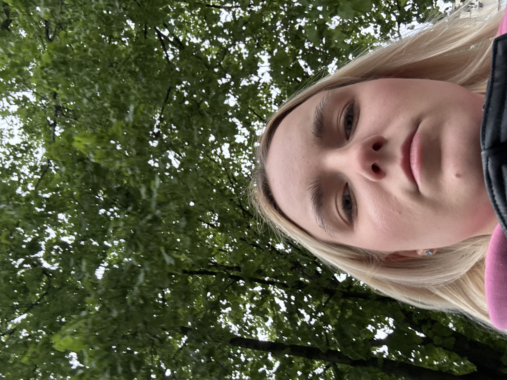 A selfie taken from a low angle shows a blonde woman against a backdrop of green foliage and sky. The woman's face is the primary focus, with her eyes, nose, and lips clearly visible. The background appears to be trees with dense leaves, possibly in a park or wooded area. The location is specified as "Unknown, Russia," but no further details about the environment or specific urban elements are discernible from the image.