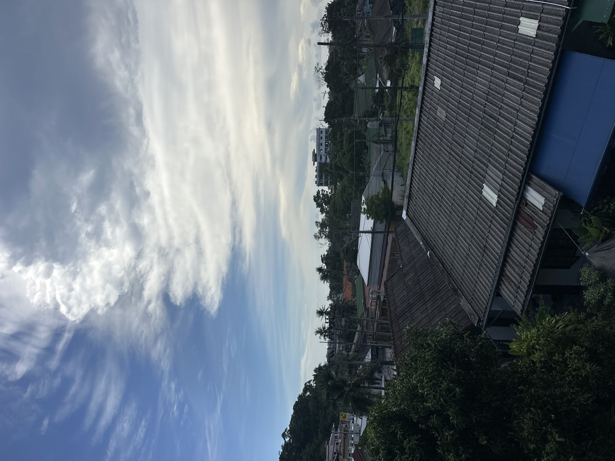This image captures a panoramic view of Karon, Thailand, under a cloudy sky. In the foreground, several tiled roofs of residential buildings are visible, some with what appear to be skylights. A large green tree on the left partially obscures some of the houses. Beyond the immediate residential area, the landscape transitions to a lush green hillside dotted with palm trees and other tropical vegetation. Further in the distance, more houses and buildings are scattered amongst the trees, with a larger, multi-story white building situated on a hill. The sky is a dramatic mix of blue patches and heavy, grey clouds, suggesting a dynamic weather pattern. The scene depicts a typical residential neighborhood in a tropical coastal town.