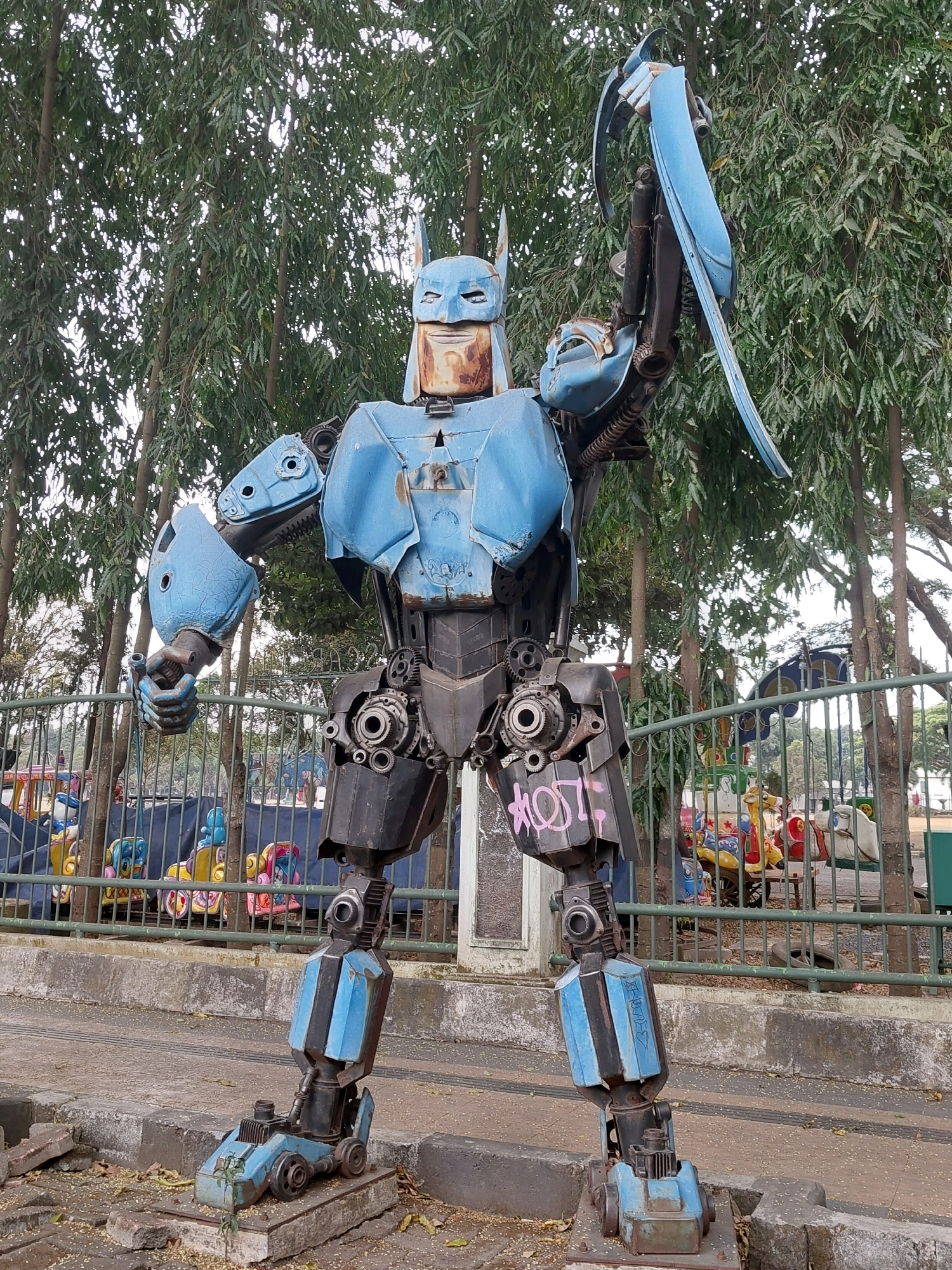 This image depicts a large, handmade robot sculpture, designed to resemble Batman, standing outdoors in a public area. The robot is constructed from various scrap metal parts, including blue painted panels, gears, and pipes. It has a distinctive cowl and cape, and its body is a mix of metallic blue and dark gray. The robot is posed with its right arm raised, holding a curved piece of metal.
The robot is situated in what appears to be a park or a public space with a fence behind it. Beyond the fence, there are colorful playground rides, suggesting the location is near an amusement area or a children's play zone. Trees with green foliage are visible in the background, indicating an outdoor setting. The ground in front of the robot is paved, with some debris and fallen leaves.
The context provided specifies that the image is from Malang, Indonesia. This type of large-scale scrap metal art is not uncommon in public spaces in some Indonesian cities, often serving as a unique form of urban decoration and a nod to popular culture. The graffiti visible on the robot's leg, with pink spray paint, suggests it is in a publicly accessible area where such markings might occur. The overall scene is one of a quirky, artistic installation in an urban environment.