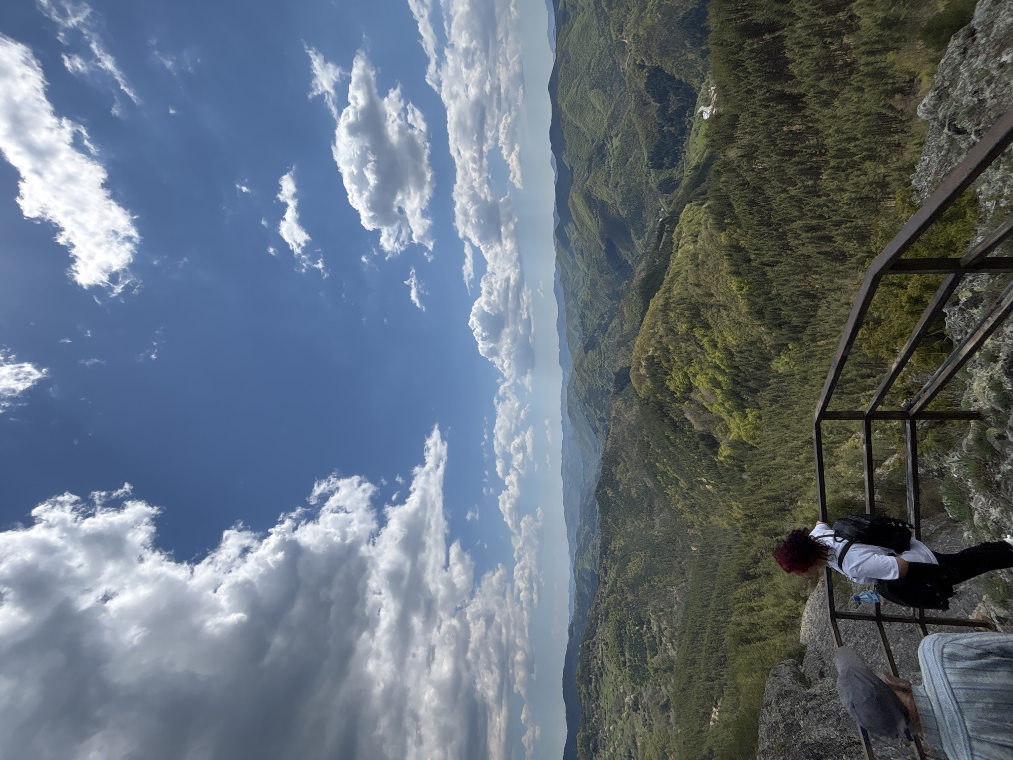 A person stands on a rocky outcrop behind a metal safety railing, gazing down at a lush, green valley. The individual is wearing a white shirt, dark pants, and a backpack. The sky above is bright blue with scattered white clouds. This image likely depicts the viewpoint at "The Eye," located in Gradat, Bulgaria. The railing is positioned at the edge of a steep cliff, providing panoramic views of the forested landscape.