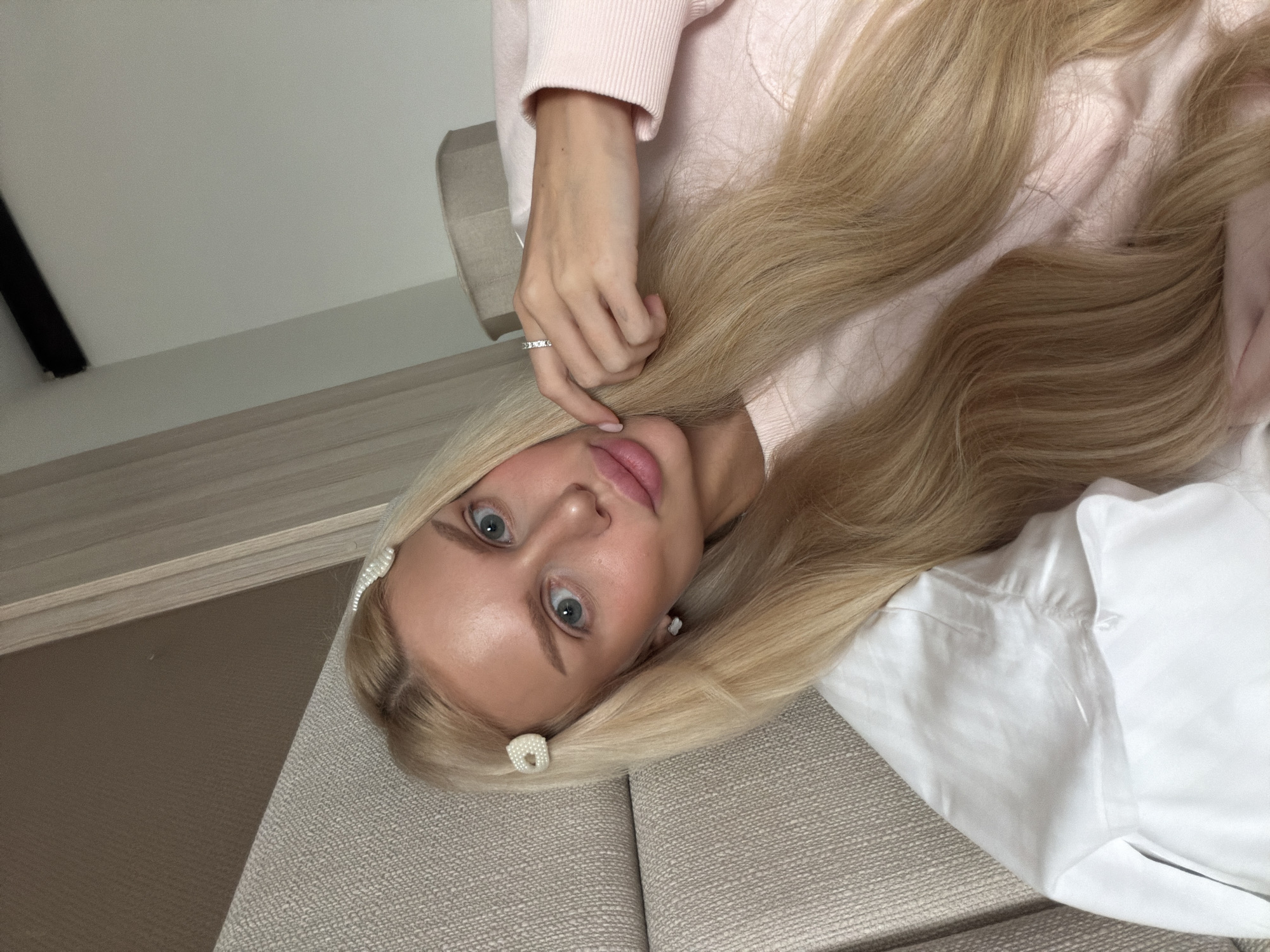 A fair-skinned woman with blonde hair lies on a beige upholstered surface, possibly a sofa. She wears a light pink sweater and a white shirt. Her blonde hair is styled with pearl-like hair clips. She has light blue eyes and wears a ring on her finger. The background includes a light-colored wall and a wooden surface. There is no evidence indicating the location is Sarajevo, Bosnia and Herzegovina.