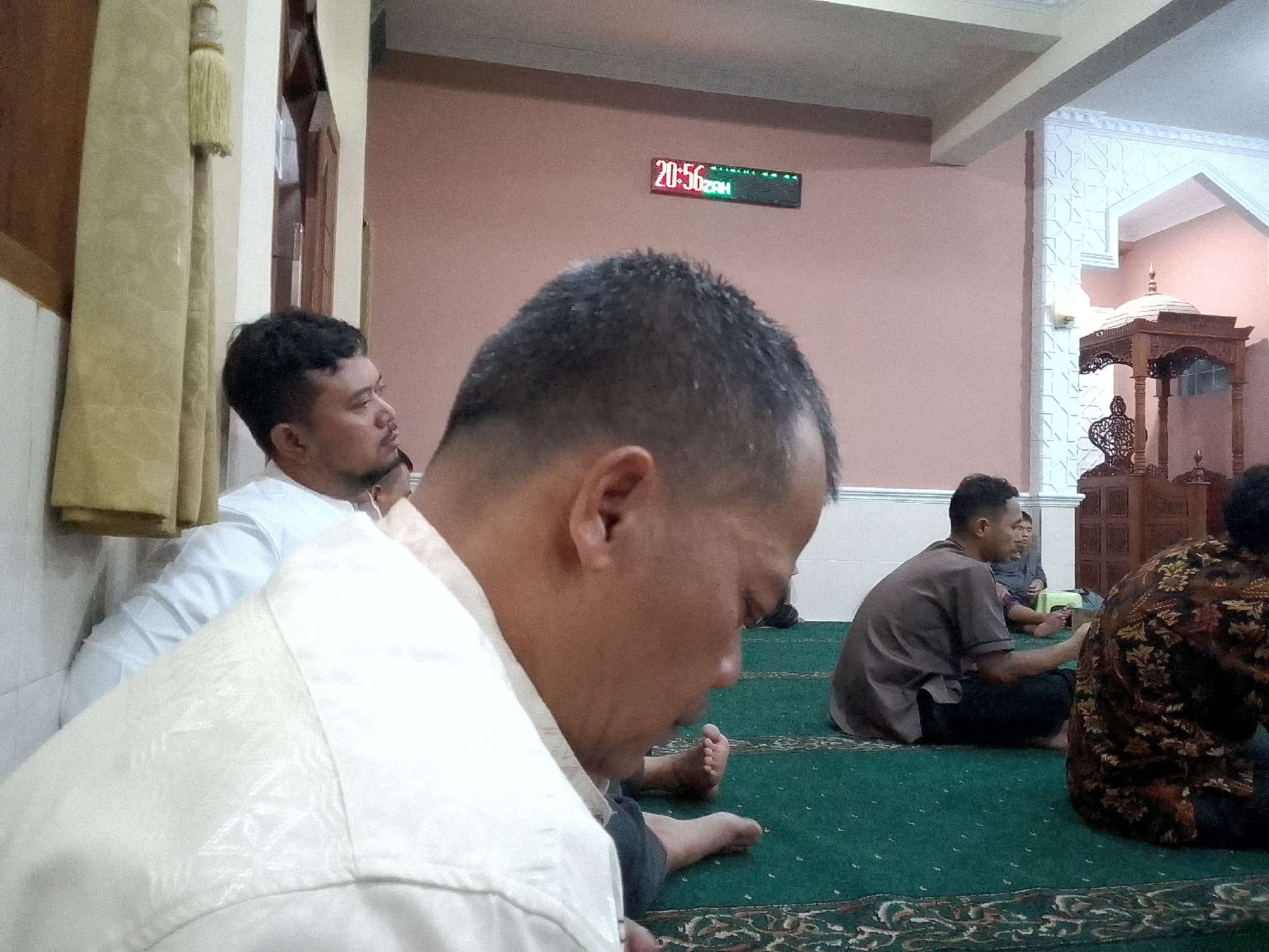 In Kertajaya, Indonesia, a group of men are gathered inside a mosque. The image captures a close-up of a man in the foreground wearing a light-colored, patterned shirt. He is seated on a green carpet with intricate patterns and appears to be looking down. To his left, another man in a white shirt is visible, also seated. In the background, several other men are seated on the carpet. One man wears a brown shirt and dark pants, while another is partially visible wearing a dark, patterned garment. A digital clock on the pink wall displays the time as 20:56. A decorative wooden structure, possibly a minbar or pulpit, is visible on the right side of the image, with intricate carvings. The environment suggests a serene and spiritual setting.
