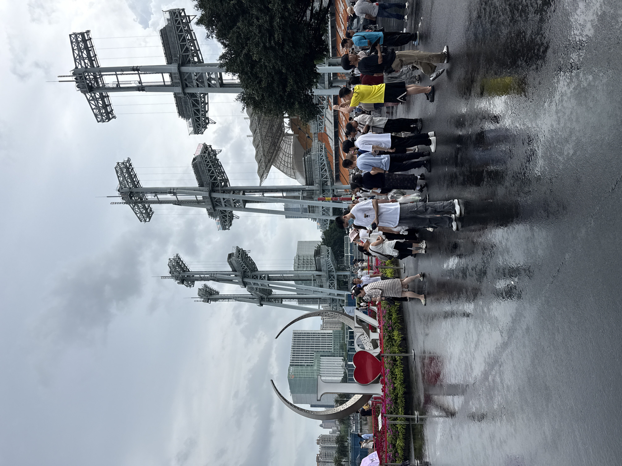 The image depicts an outdoor scene on a cloudy day, with a wet, reflective ground surface, indicating recent or ongoing rain. A group of people are gathered in front of a large, decorative sculpture that spells out "I LOVE GZ", with a red heart as the "O". Behind the sculpture, a stadium with orange seating is visible, flanked by tall, industrial-looking structures with lights. Buildings are visible in the background. The overall atmosphere suggests a public gathering or event. The location is identified as Tianhe District, China.