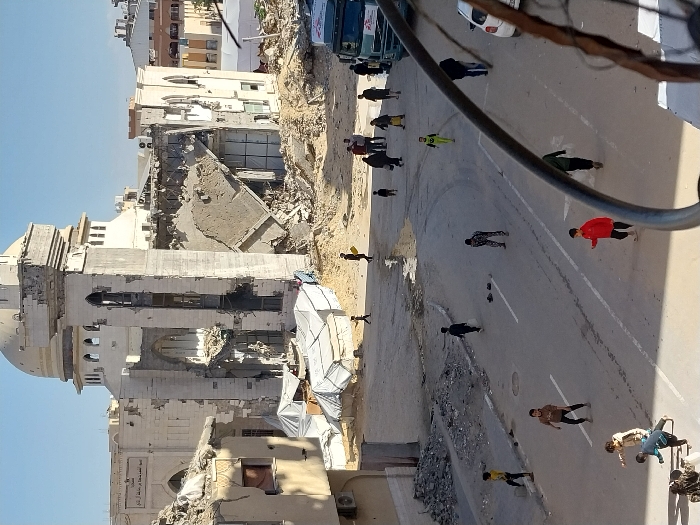 The image depicts a severely damaged urban landscape in Khan Yunis, Palestinian Territory, under a clear sky. In the background, the heavily damaged Al-Faraheen Mosque stands prominent, featuring a distinctive dome and a partially collapsed minaret-like tower. Large sections of its structure are missing, exposing interior elements and extensive debris. Adjacent buildings are also significantly compromised, with collapsed walls and rubble scattered throughout the scene.
In the mid-ground, multiple white tarpaulin tents, some appearing to serve as makeshift shelters, are set up amidst the debris piles. One tent displays a visible red logo, indicative of Médecins Sans Frontières (MSF). A wide, paved street with white lane markings runs from the mid-ground into the foreground, showing patches of dirt and a noticeable puddle on its surface.
The street is populated by individuals. Approximately ten children are scattered across the foreground and mid-ground of the road, many engaged in play, with some running or interacting. Several adults are walking on the right side of the street, near a parked white and green truck. A white car is partially visible on the far right. The overall environment conveys widespread structural damage and ongoing daily life amidst the destruction.