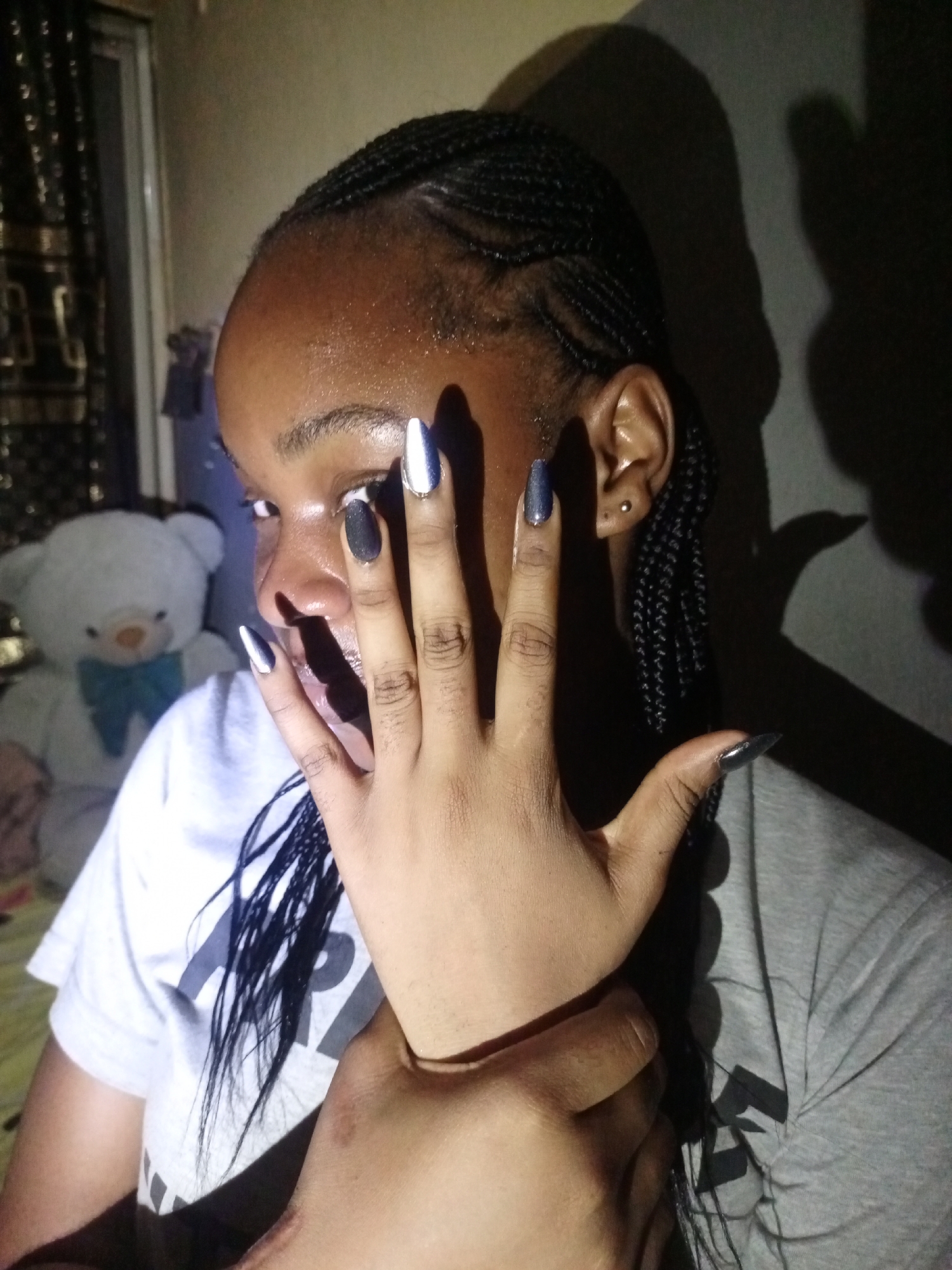 A young Black woman is indoors, partially obscured by her own hand, with a background featuring a teddy bear, curtains, and a wall casting shadows. She is wearing braids. Her fingernails are painted in varying shades of gray and silver. Her facial expression is one of slight surprise, with her mouth open. The lighting is dim. There is no way to verify the exact location within Keffi, Nigeria based on this image alone.