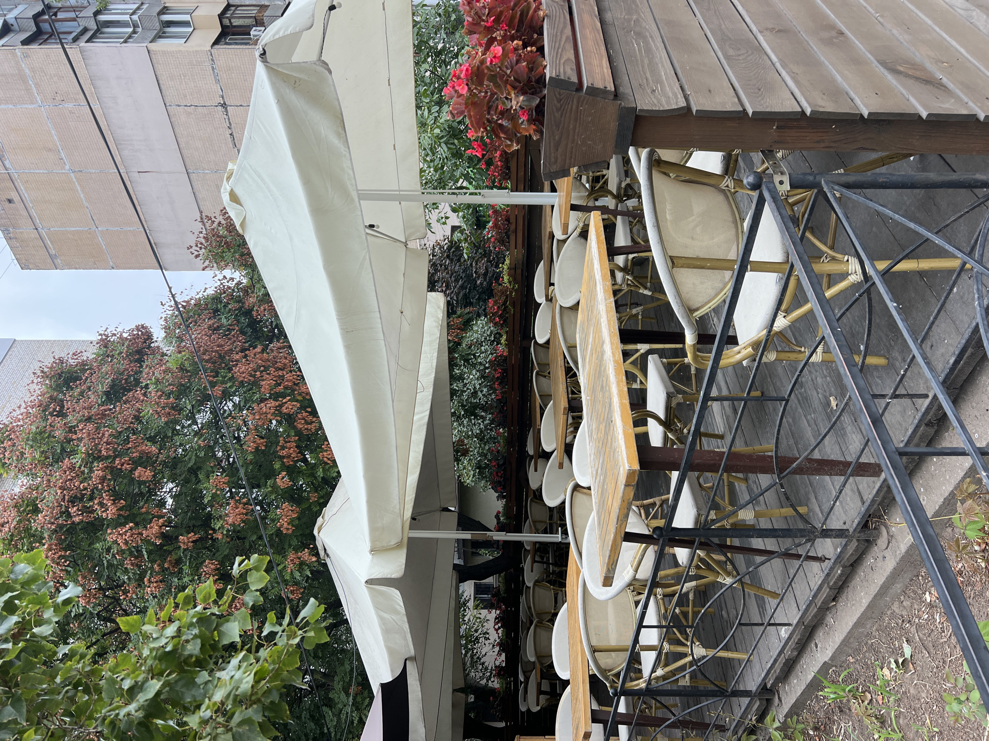The image shows an outdoor seating area of a cafe or restaurant, likely located in Odesa, Ukraine. The scene is set on a wooden deck with multiple tables and chairs covered by large white umbrellas. The tables are made of light-colored wood, and the chairs appear to be made of wicker or a similar material with light-colored cushions. A black metal railing encloses the seating area, separating it from a small patch of ground with some greenery. In the background, there are buildings, one of which has a distinctive tiled facade, and mature trees with red foliage. The sky is overcast. There are no people visible in the image. The overall impression is one of a quiet, possibly slightly disused, outdoor dining space.