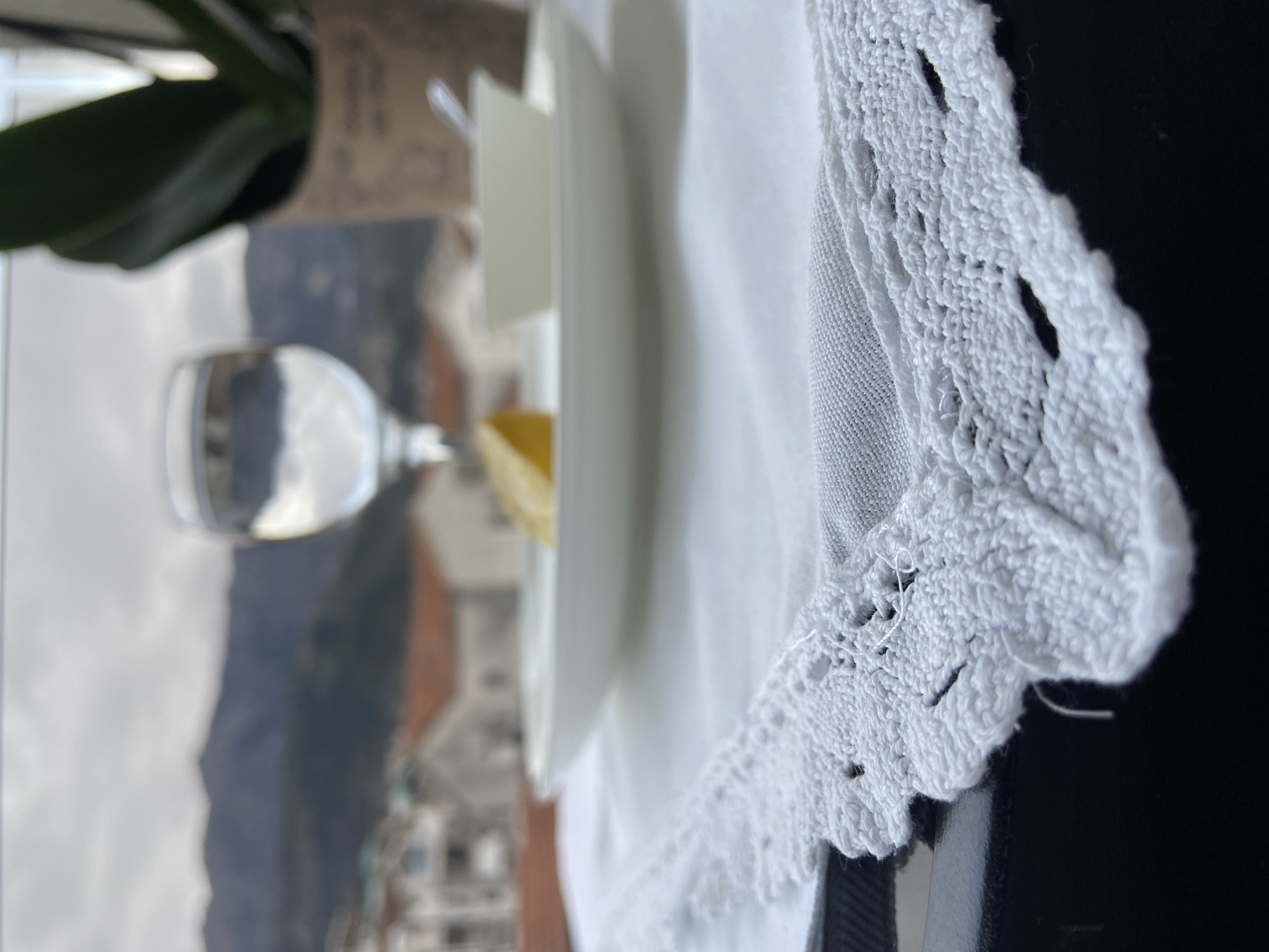 Here is a detailed description of the image:
The image depicts a table setting with a glass of water, plates, and a white cloth with a lace edge. In the background, out of focus, is a cityscape with mountains.
Based on the architecture and the landscape, it's very likely the location is Budva, Montenegro. The buildings in the background appear to be the typical red-roofed structures found in Budva's old town and the mountainous backdrop is consistent with the area.