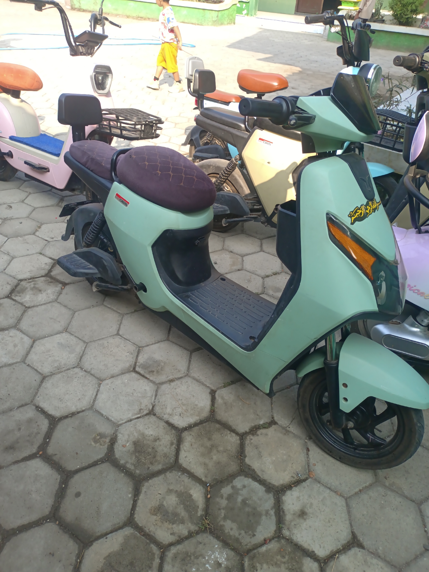 In Sarimulyo, Indonesia, a mint green electric scooter with a dark patterned seat and a small black windshield is parked on a hexagonal paver patio. The scooter has a black floorboard and orange turn signals. To the left, a pink and beige scooter with a blue seat pad is visible. In the background, a person wearing a white t-shirt with a colorful graphic and yellow shorts walks away from the scooters. The environment appears to be outdoors with some vegetation present.