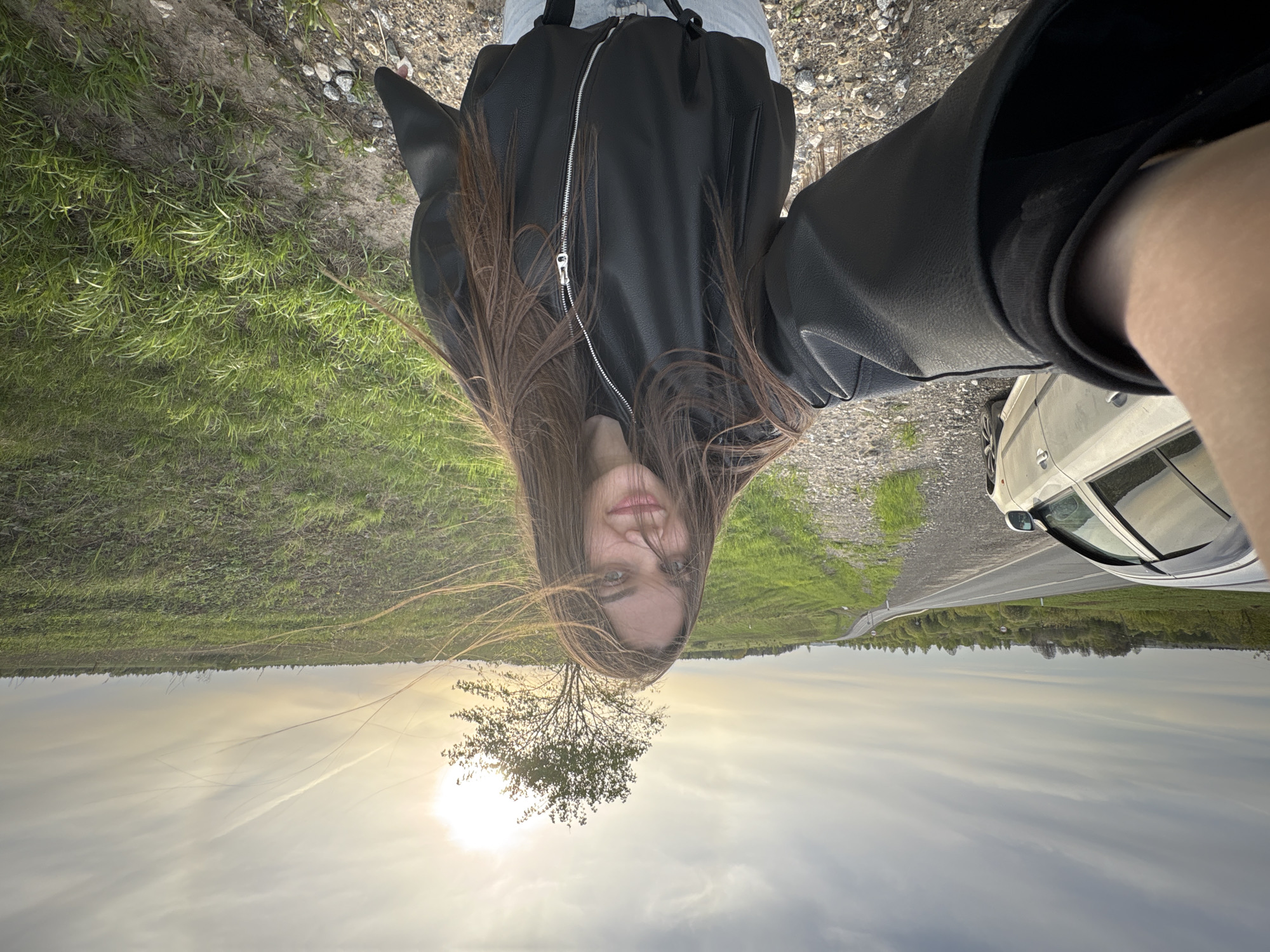 A woman stands near a car on a road in a rural area of Бикбарда, Russia, during the daytime. She faces the camera wearing a black leather jacket and jeans. The location appears to be a roadside with grass fields and a solitary tree between the road and sky. The sky is visible, with some cloud cover and bright sunlight.