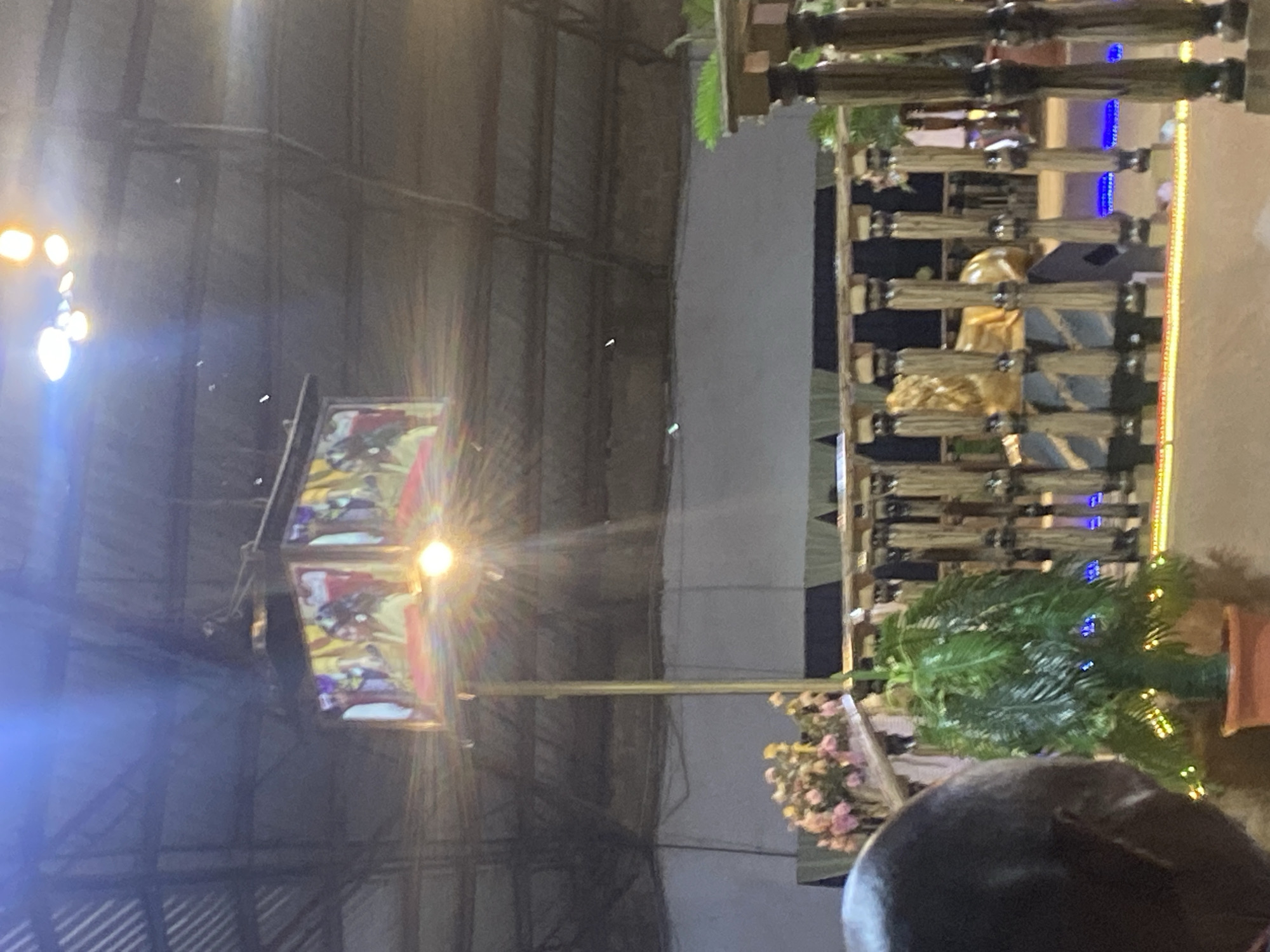 An indoor event space, likely a hall or a tent, with a corrugated metal roof. A large, suspended screen displays an image of people, possibly performers or a religious depiction, illuminated by a bright light bulb hanging directly below it. In the foreground, a person's head and shoulder are visible in profile, facing away from the camera. To the right of the person, a decorative railing with carved wooden posts and a golden lion statue is present. A potted plant with green foliage and colorful lights sits near the railing. The background features a white wall or curtain. The overall scene suggests a lively event, potentially a church service or a celebration, taking place in Rukuba Road, Nigeria.