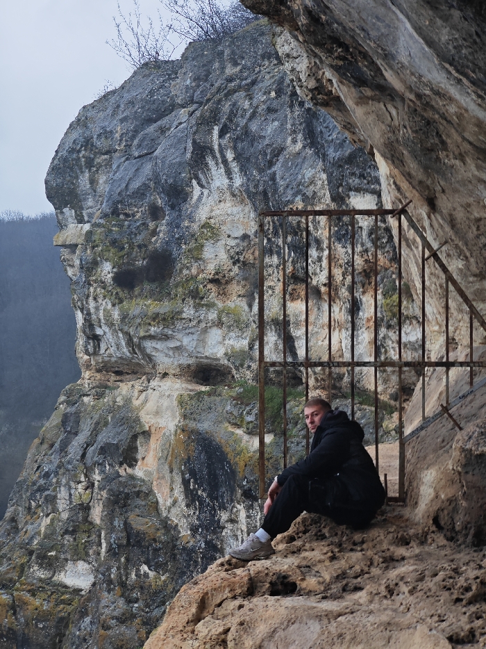 A male individual is seated on a textured rock ledge within a natural rock formation, characteristic of the Shumen Plateau near Shumen, Bulgaria. He is positioned towards the right of the frame, facing forward with a direct gaze towards the camera. He wears a dark-colored puffer jacket, dark trousers, and light grey athletic shoes. His right knee is bent, while his left leg extends forward. Behind him and to his left, a rusty, dark-colored metal gate with vertical bars and angled supports is affixed to the rock face, enclosing a section of the rock overhang or cave entrance. The surrounding rock formations exhibit varied shades of grey, white, and ochre, with visible stratification, erosion patterns, and scattered patches of green moss. In the distant background, a prominent, rugged cliff face looms to the left, overlooking a hazy, densely forested valley under an overcast sky. Minimal, bare tree branches are visible atop the furthest rock peak.