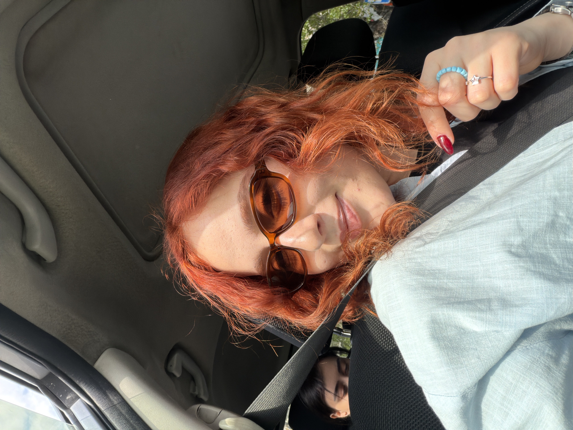 An eye-level selfie captures a person with vibrant red, wavy hair and brown-framed sunglasses pushed onto their face, taken inside a car during the daytime. The individual, likely female, is looking towards the camera with a subtle smile.
They are wearing a light blue, possibly linen, shirt over a darker garment. Their right hand is visible, adorned with a light blue beaded ring on the middle finger and a silver star-shaped ring on the ring finger. Long, pointed nails painted in dark red are also prominent. A silver watch is partially visible on the left wrist.
The interior of the car is visible, including the light grey headliner and the seatbelt that diagonally crosses the frame. Bright sunlight illuminates the scene, creating highlights on the person's hair and face. Through the car windows, out-of-focus green foliage suggests an outdoor environment. A faint reflection of another person with dark hair and sunglasses can be seen in the lower part of the image, possibly in a car mirror or a reflective surface. No visible text is present in the image.