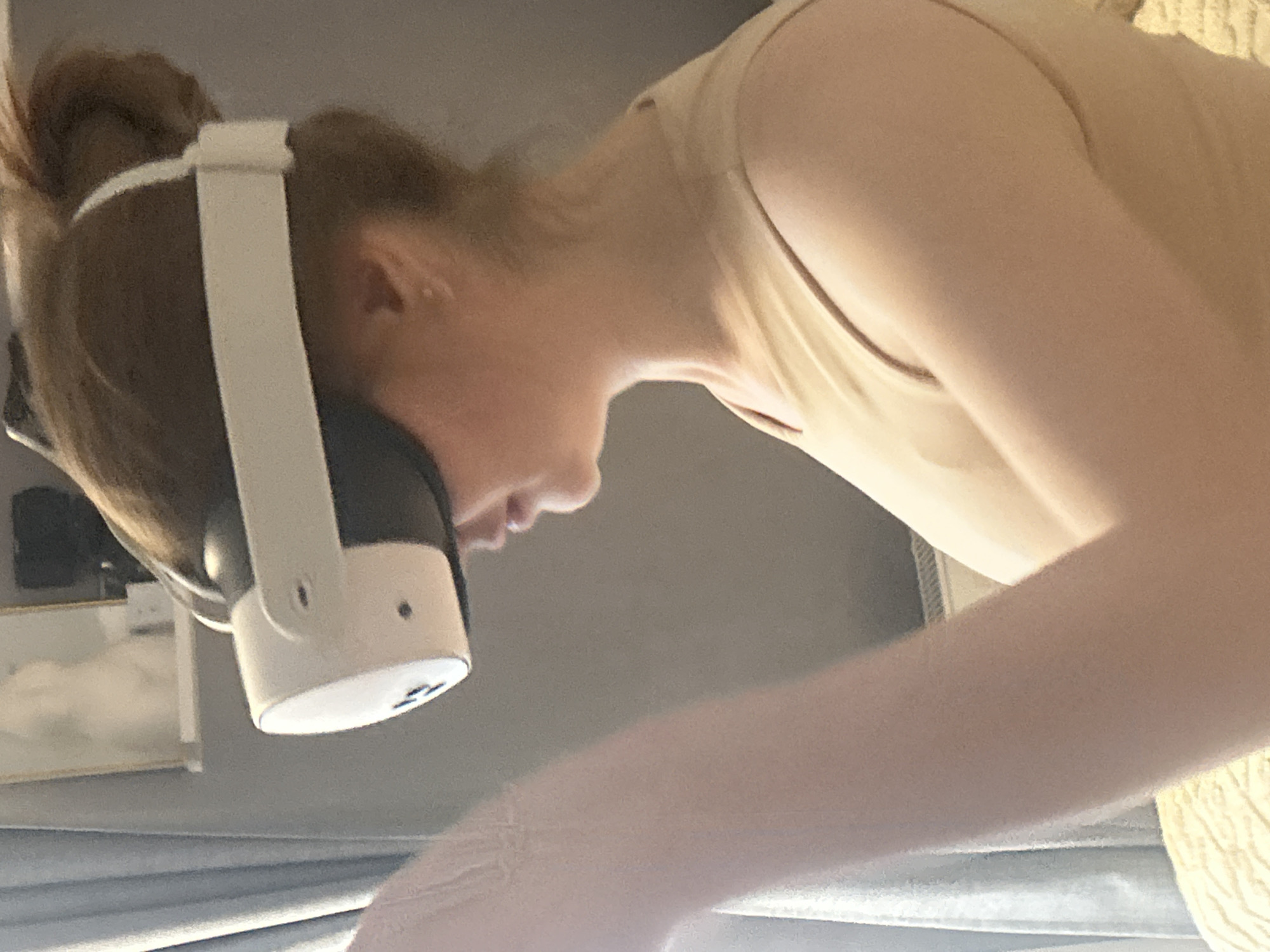 The media file shows a person wearing a VR headset and a light-colored tank top. The person's head is bowed down, and their hair is pulled up. The VR headset is white with black accents. The background is a muted gray wall with a hint of a patterned surface on the right side, possibly a couch or bed. A shelf with framed pictures or artwork is visible on the left. The lighting suggests an indoor setting, likely during the day. The image is oriented sideways, with the person's head pointing towards the bottom of the frame. The person's facial features are partially obscured by the headset, but their profile is visible. The overall scene conveys a sense of engagement with the virtual reality experience. No visible text is present in the image.