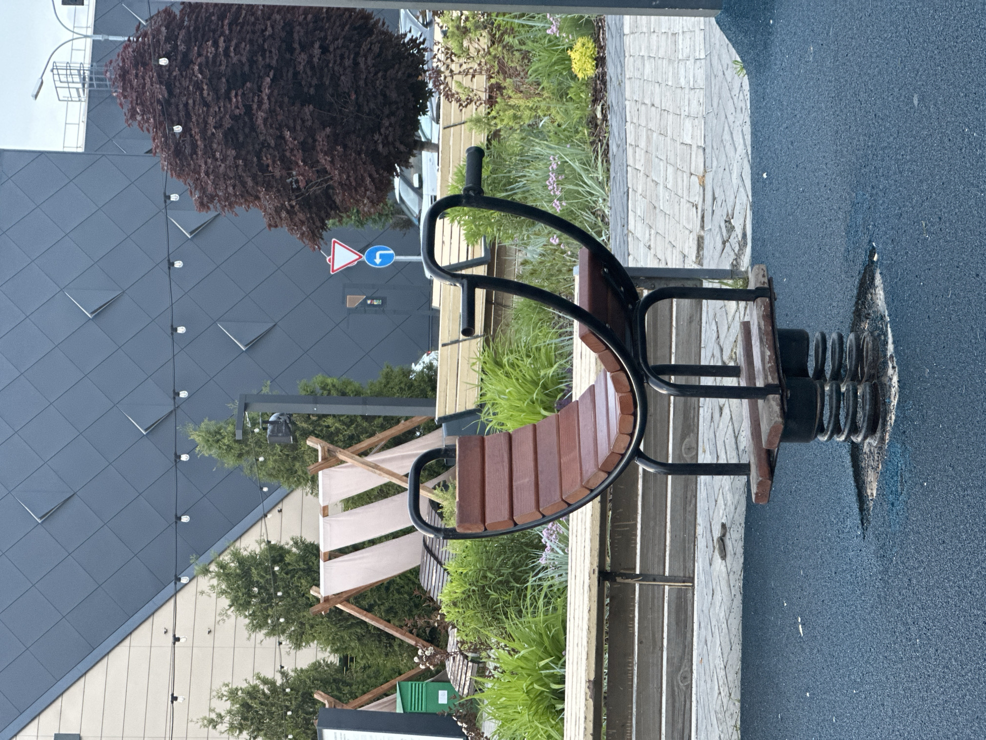 The image depicts a child's spring rocker in an outdoor playground setting. The rocker has two seats, each with a wooden back and seat, and black metal frames with hand grips. It is mounted on a large black spring attached to a base embedded in a dark blue, textured playground surface.
Behind the rocker, there is a landscaped area with various plants and shrubs. A wooden bench runs along the back of the planter. In the background, there's a building with dark grey, triangular patterned siding and a large dark red tree.
A street sign with a blue left turn arrow and a red and white triangular yield sign is visible near the tree. Further back, a structure with a white awning supported by wooden posts is present. The lighting suggests it is daytime.
Based on the style of playground equipment and the surrounding architecture, the location is likely Kazan, Russia.