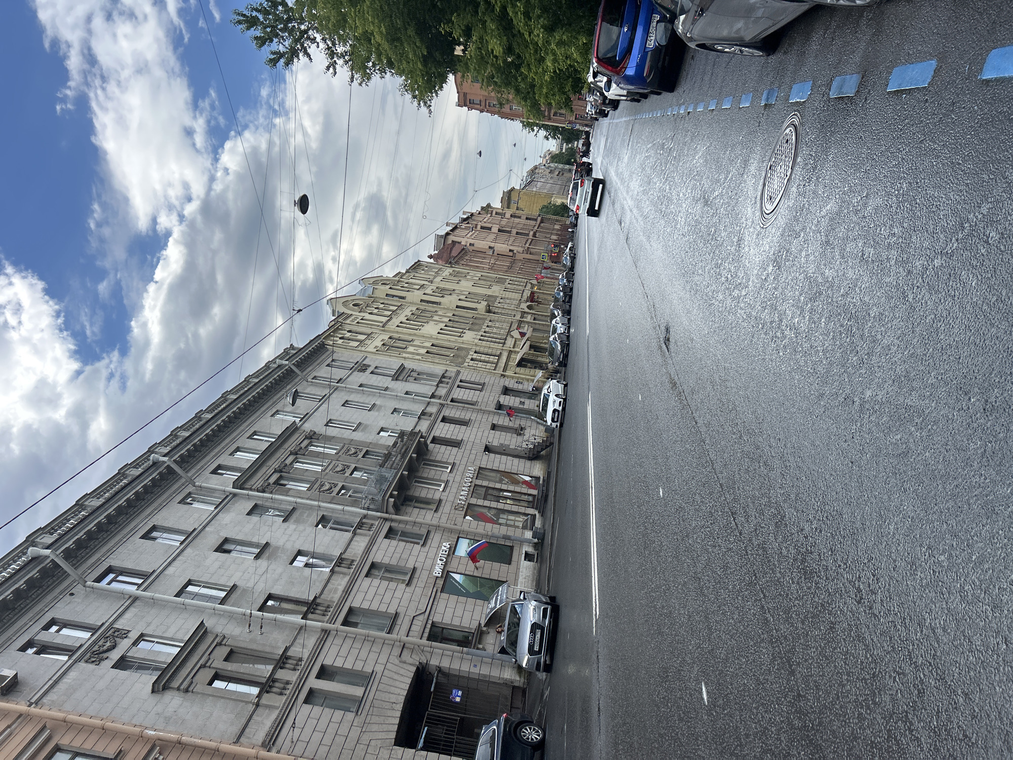 The image depicts a street scene in an urban environment. The perspective is from street level, looking down a wide, wet asphalt road. On the left side of the road, a row of multi-story buildings with ornate architectural details lines the street. These buildings appear to be older, with many windows and balconies. Several cars are parked on the street and some are in motion. On the right side of the road, there are more buildings, one appearing to be a brick structure, and a line of parked cars. The sky is partly cloudy with patches of blue sky visible. The scene suggests a bustling city with ongoing activity.