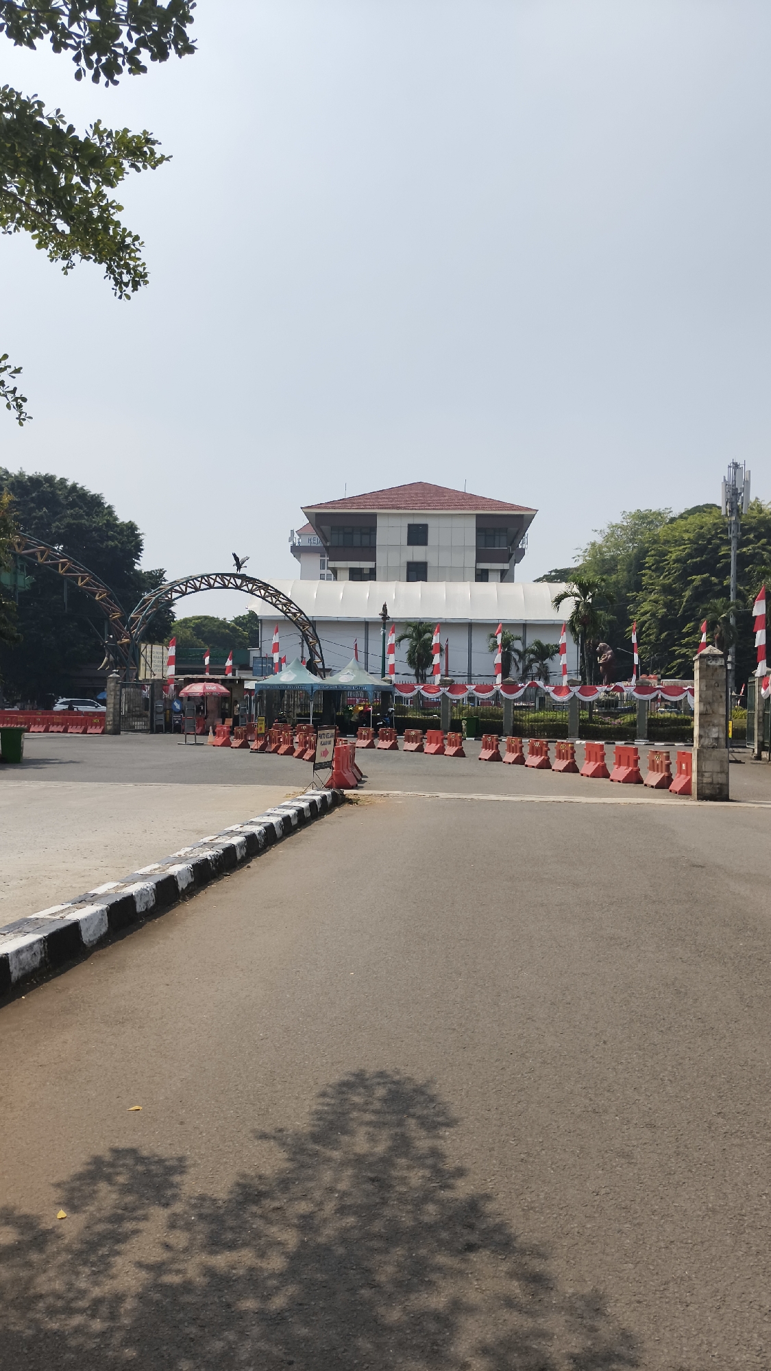 The image depicts the entrance to the National Archives Building in South Jakarta, Indonesia, on a clear, sunny day. The building, a multi-story structure with a distinctive red-tiled roof, is visible in the background. The foreground shows an asphalt road leading towards the entrance, with a black and white striped curb along the left side.
The entrance area is decorated with red and white Indonesian flags, arranged along a fence and on poles. Red and white banners are also draped across the fence. Several red traffic barriers are placed on the road, directing traffic flow. Two green tents are set up near the entrance, and a small red umbrella is visible. A large, arched metal structure with a bird sculpture on top stands at the gateway. Beyond the entrance, more trees and foliage are present, contributing to the urban green space. Cell phone towers are also visible on the right side of the image. The ground is somewhat shaded by trees, casting dappled shadows on the road in the foreground. No people are clearly discernible in the image.
