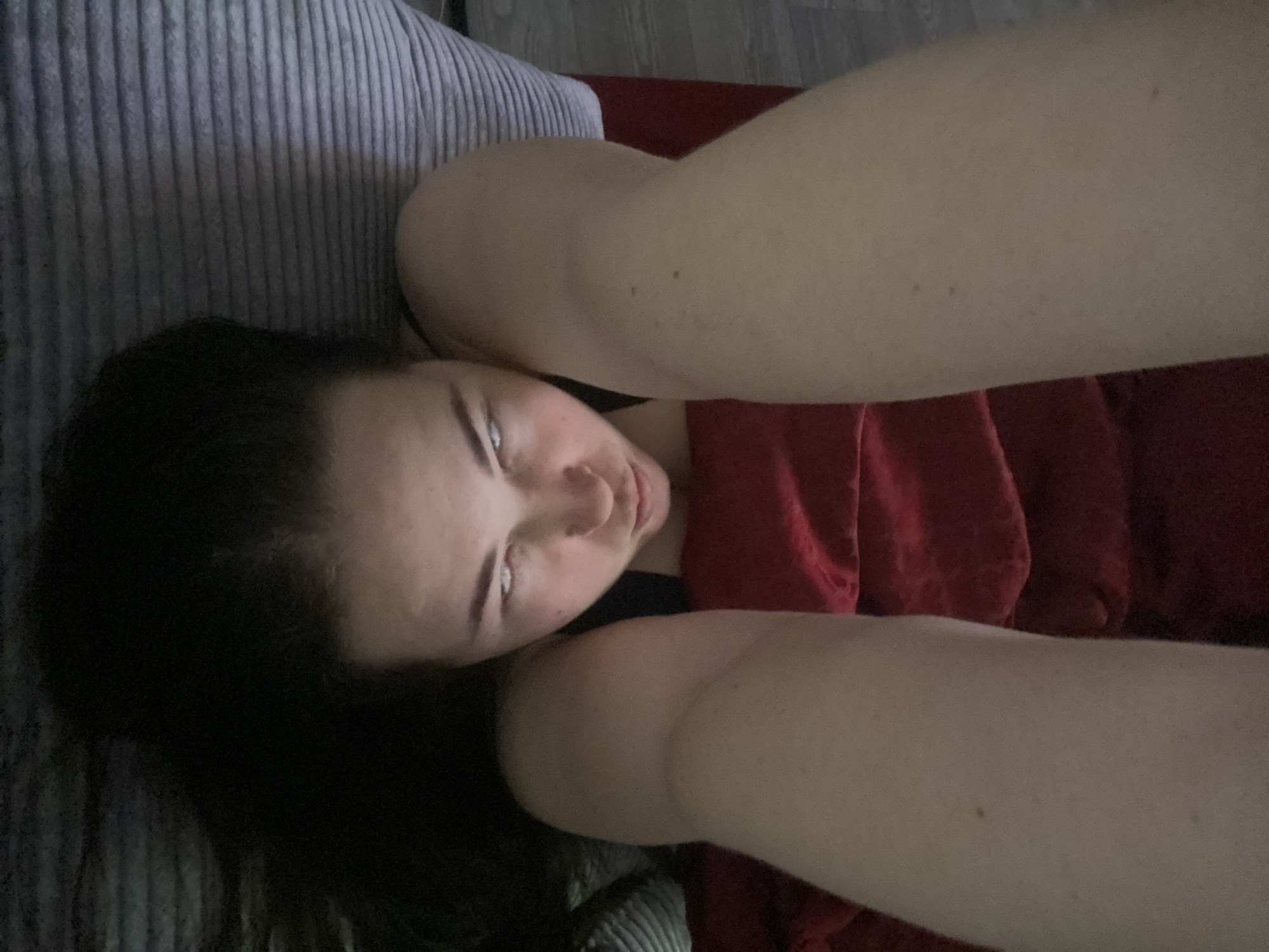 A woman lies on her back, positioned between a gray cushioned sofa and a red fabric surface. Her dark hair is spread on the cushion. She looks directly into the camera with her eyes angled upwards. Her arms are raised toward the camera, obscuring some of her body. The location is impossible to discern from the image.