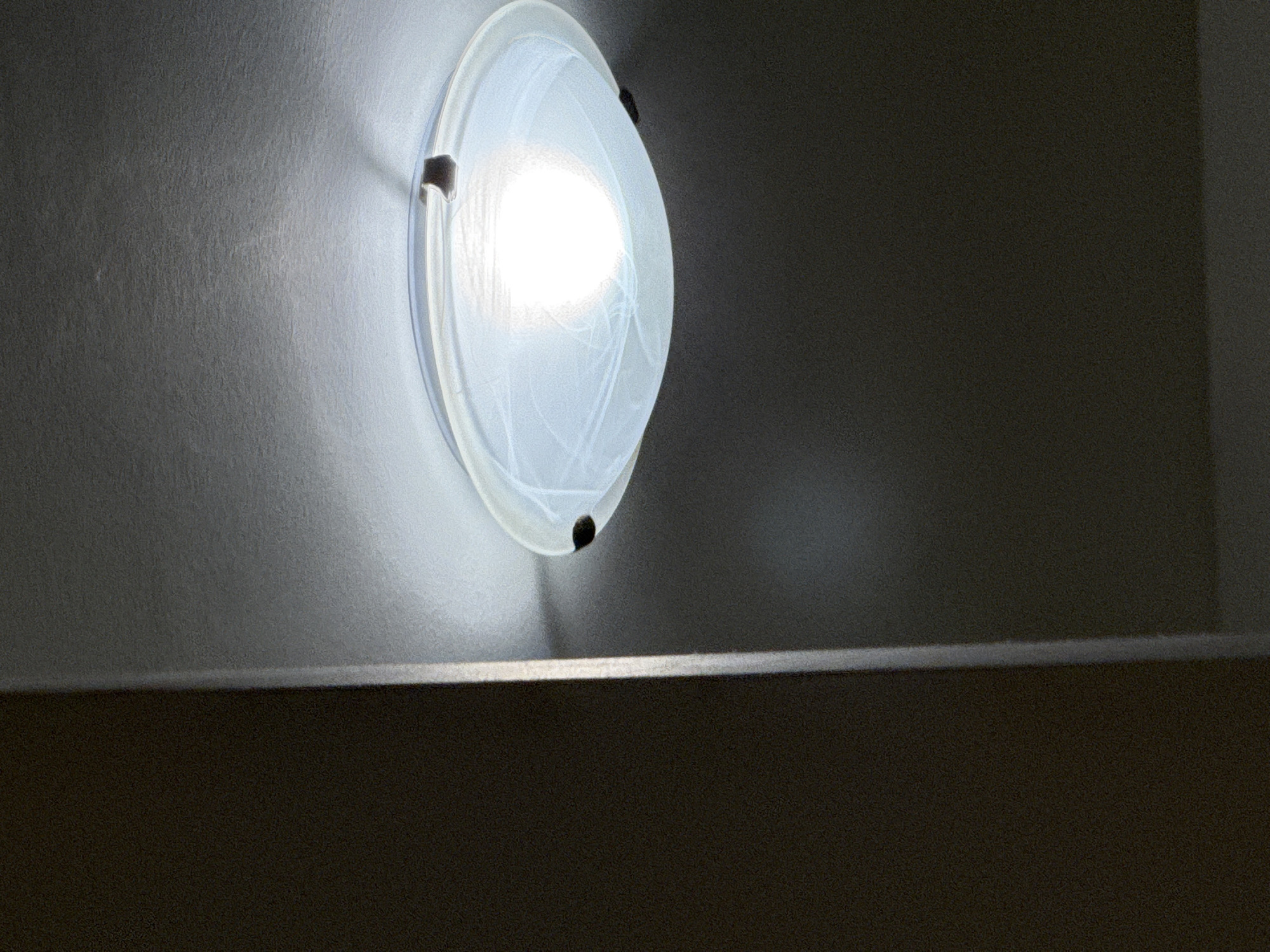 This media file shows a close-up, low-angle shot of a modern, circular wall-mounted light fixture. The fixture has a frosted glass or plastic cover with faint, swirling patterns, behind which a bright light emanates. Two dark, metallic brackets are visible, securing the fixture to a textured, light-colored wall. The wall appears to be painted or stuccoed, with subtle variations in tone and texture, suggesting it is an interior wall. To the right of the light fixture, the wall transitions to a darker, more uniform gray, indicating a change in surface or a shadowed area. In the foreground, a dark, horizontal surface is visible, creating a sharp division in the frame. This surface has a matte finish and appears to be furniture or a ledge. The lighting suggests a dimly lit interior space, possibly at night or in a room with minimal natural light. The overall impression is one of a contemporary interior design element, likely found in a hallway, staircase, or a decorative accent wall. There is no text visible in the image. The context of Budva, Montenegro, suggests this could be an interior shot within a hotel, apartment building, or a private residence in that coastal city.